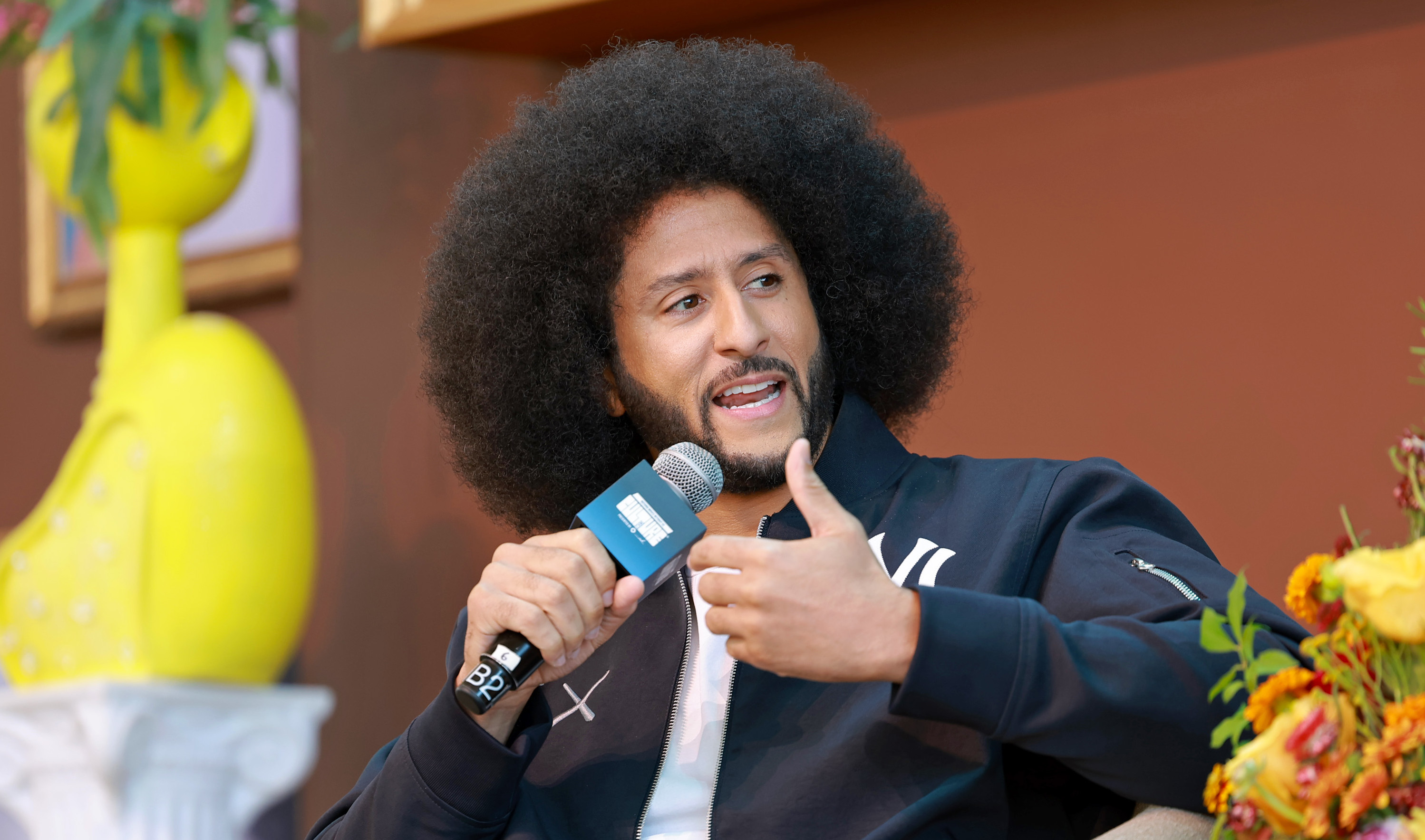 Colin Kaepernick speaking into a microphone, sitting on a chair with colorful flowers nearby.