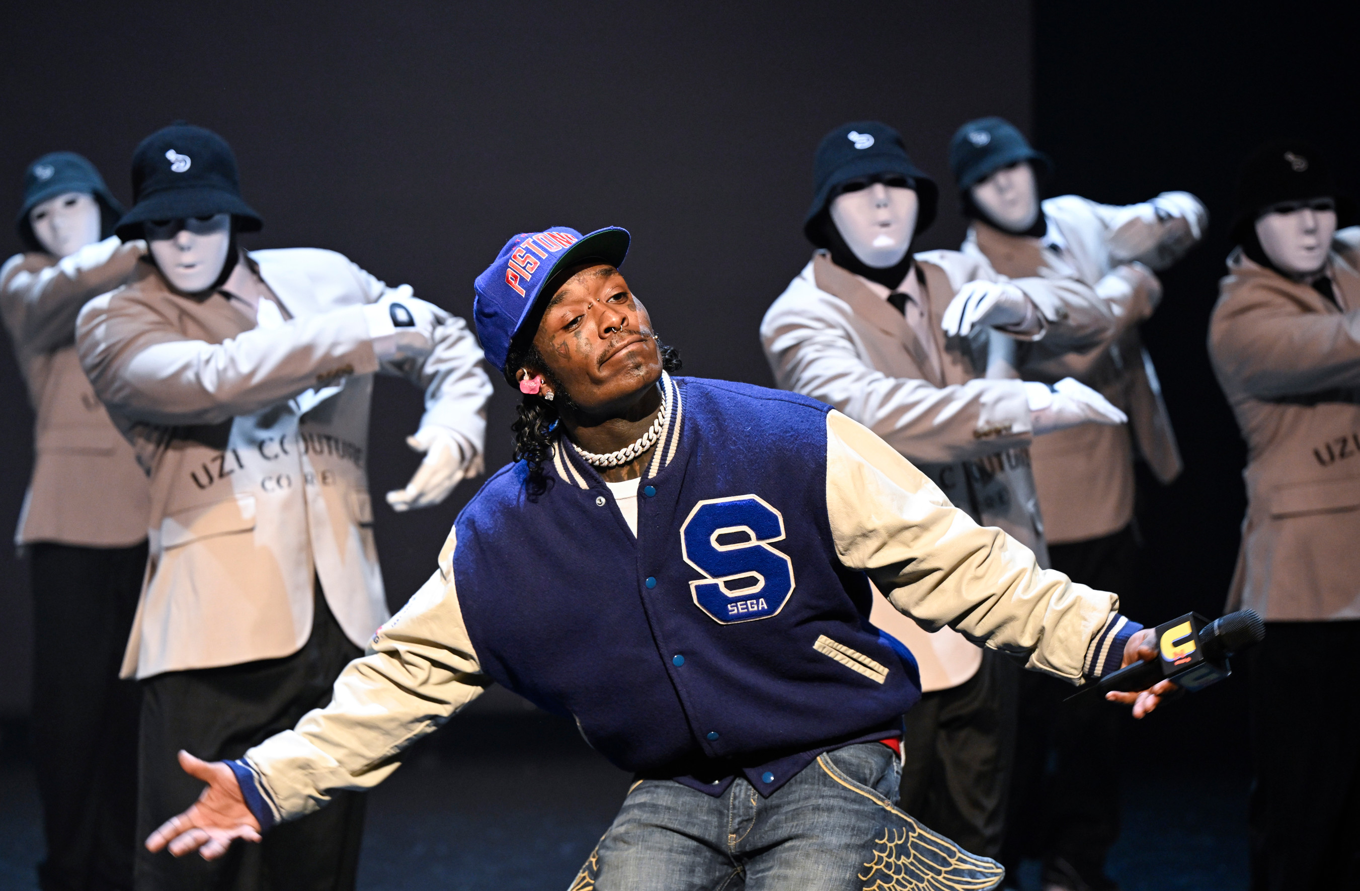 A performer in a blue varsity jacket and cap on stage, surrounded by dancers in beige jackets and white masks.