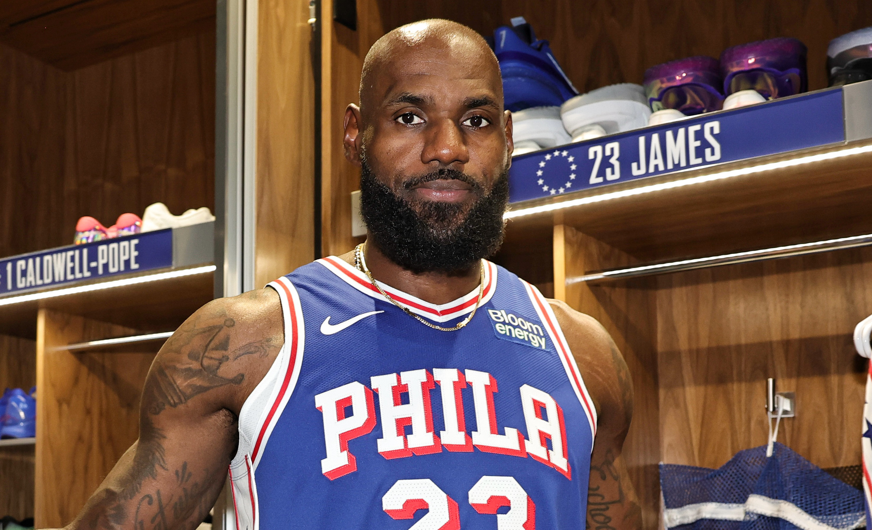 LeBron James in a Philadelphia 76ers jersey number 23 stands in a locker room, arms outstretched, with open lockers behind him.