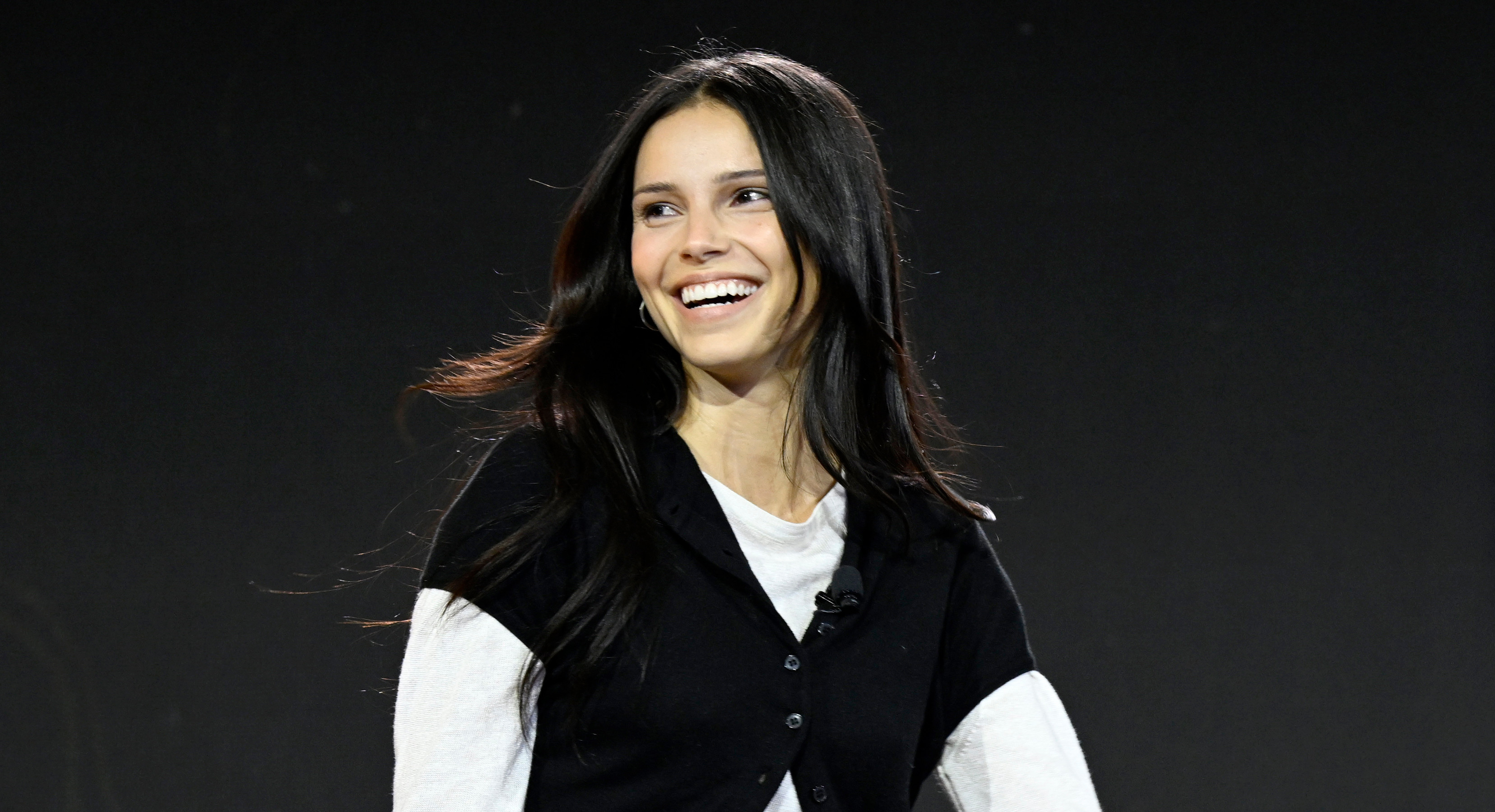 Inde Navarette with long dark hair is smiling and walking on stage, wearing a black vest over a white long-sleeve top and black pants.