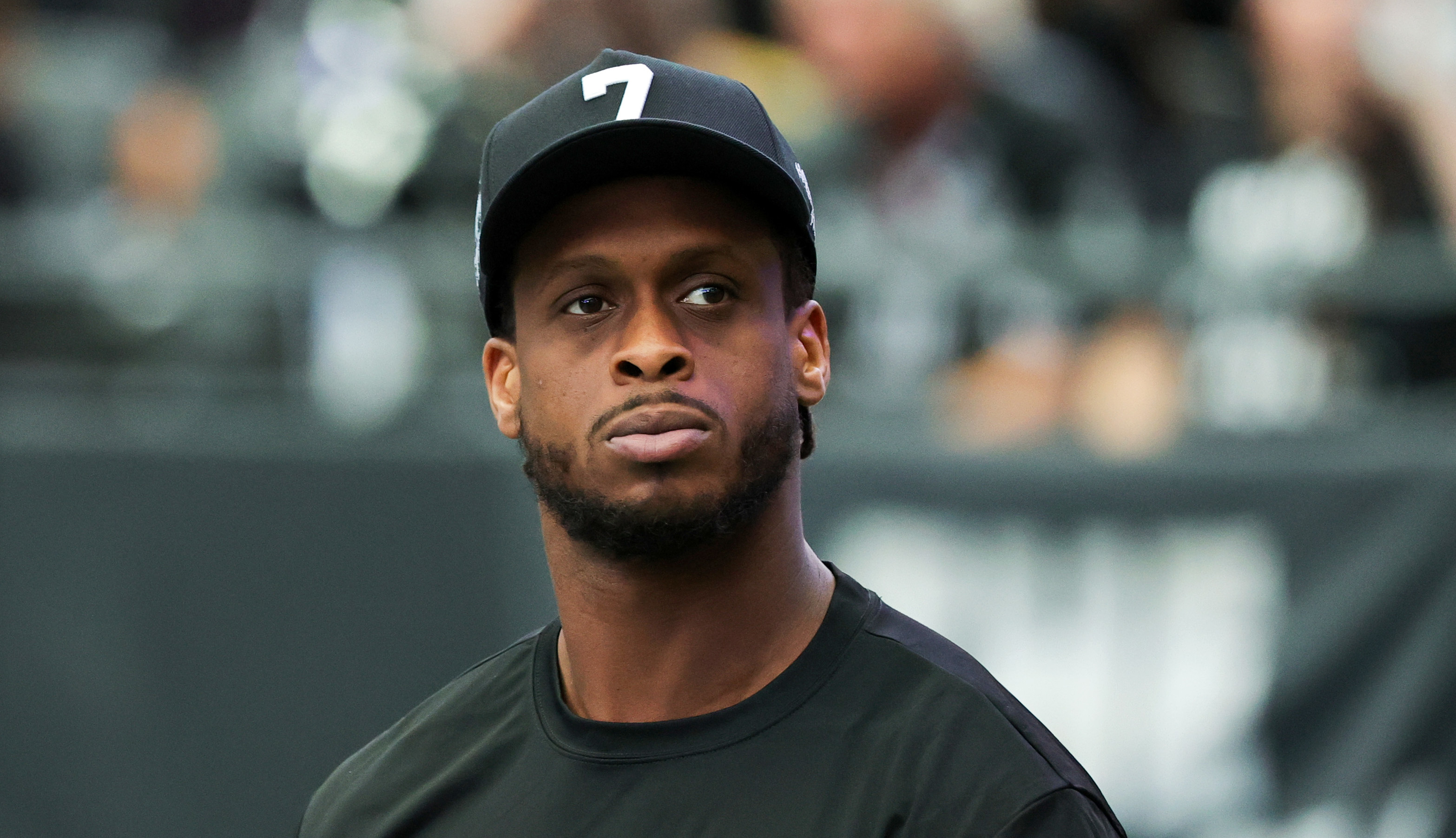Geno Smith wearing a Raiders cap and shirt stands on the field, looking focused. The background is blurred with people in the stands.