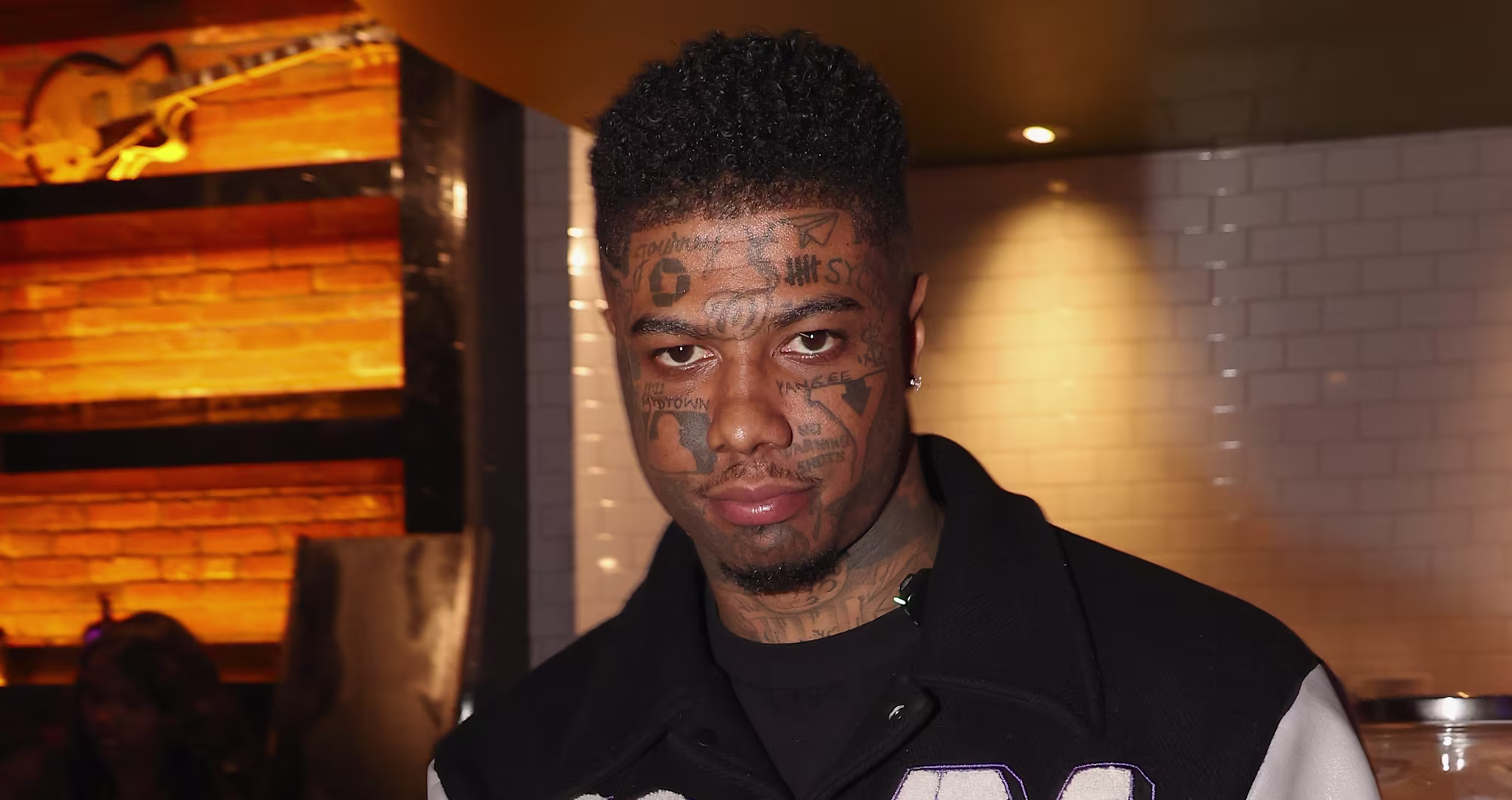 Blueface with facial tattoos and a varsity jacket poses in a restaurant, holding a phone and gesturing with his hand.
