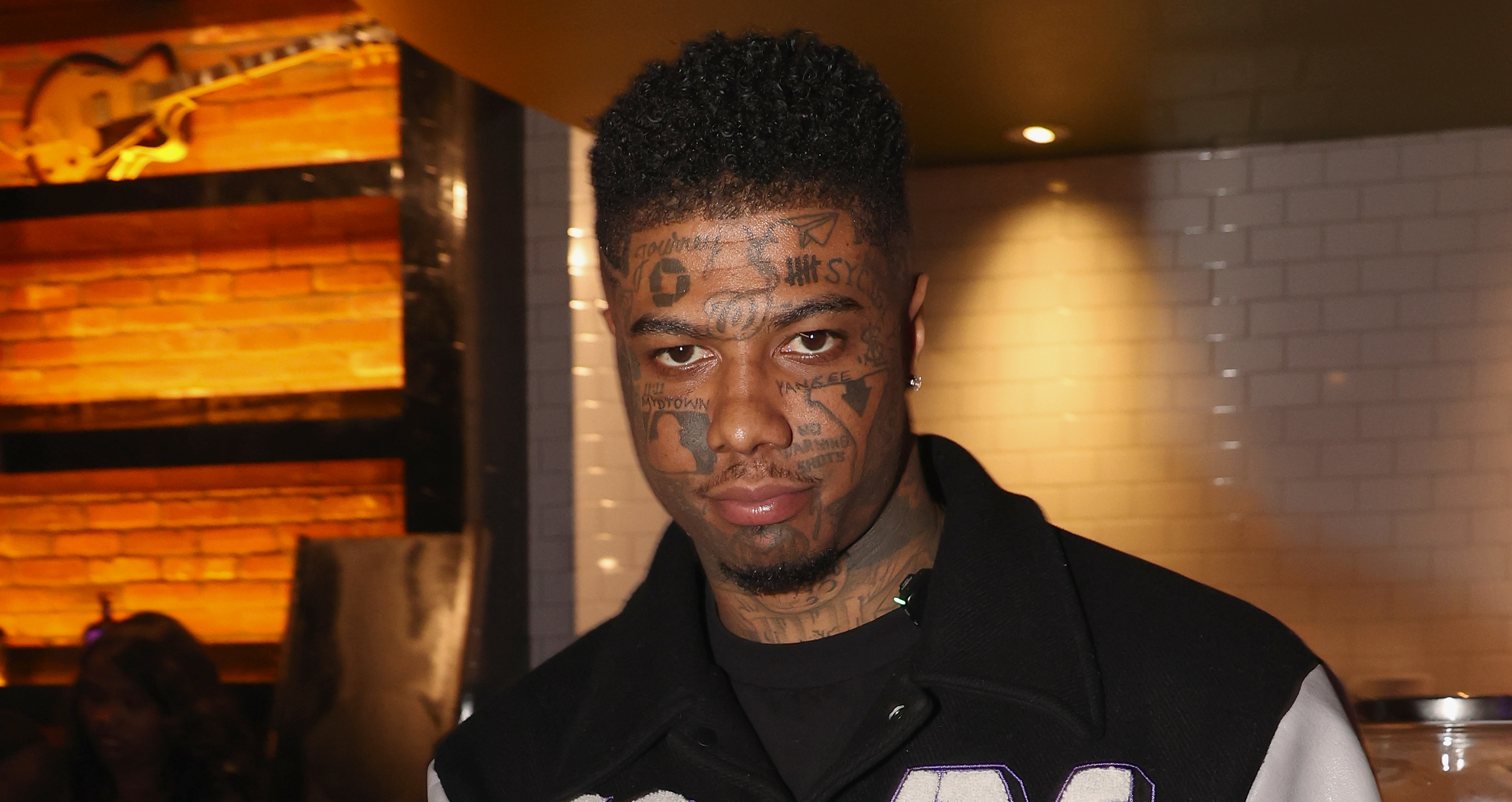 Blueface with facial tattoos wearing a black and white varsity jacket, standing in a restaurant, holding a phone, and making a hand gesture.