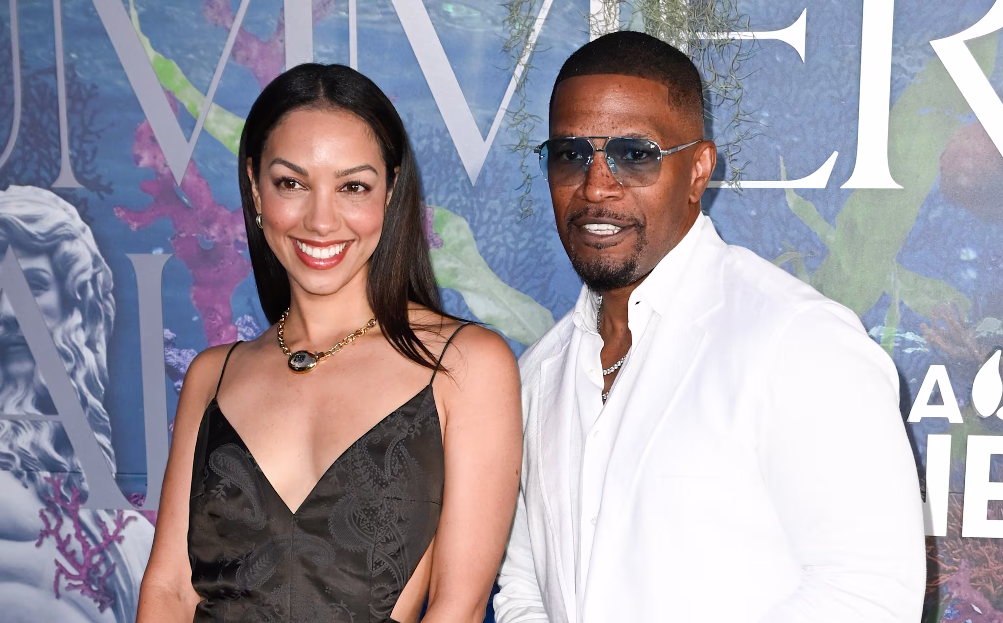 Corrinne Foxx in a black dress and Jamie Foxx in a white suit with sunglasses stand together at an event with a decorative backdrop.