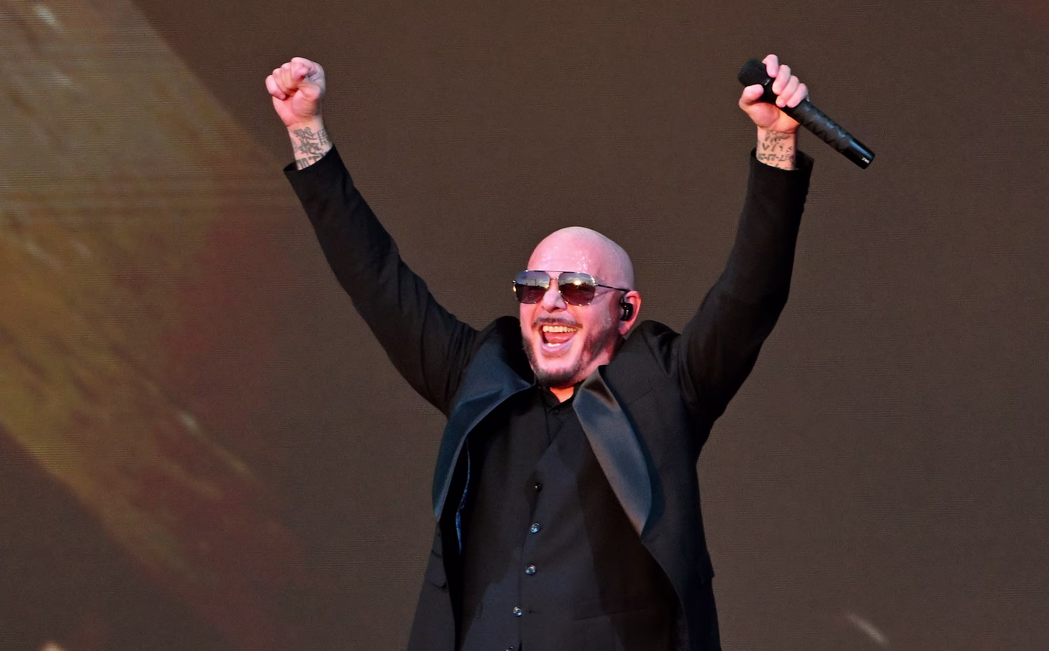 Pitbull in a black suit and sunglasses, holding a microphone, raises his arms triumphantly on stage.