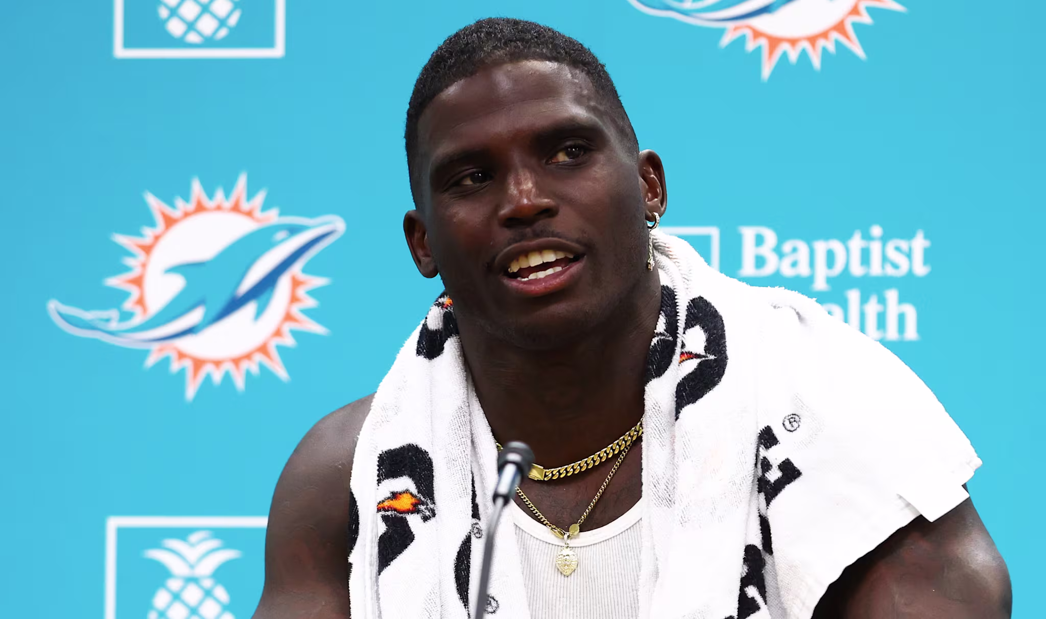 Tyreek Hill at a press conference, wearing a tank top and towel, with a Miami Dolphins backdrop.