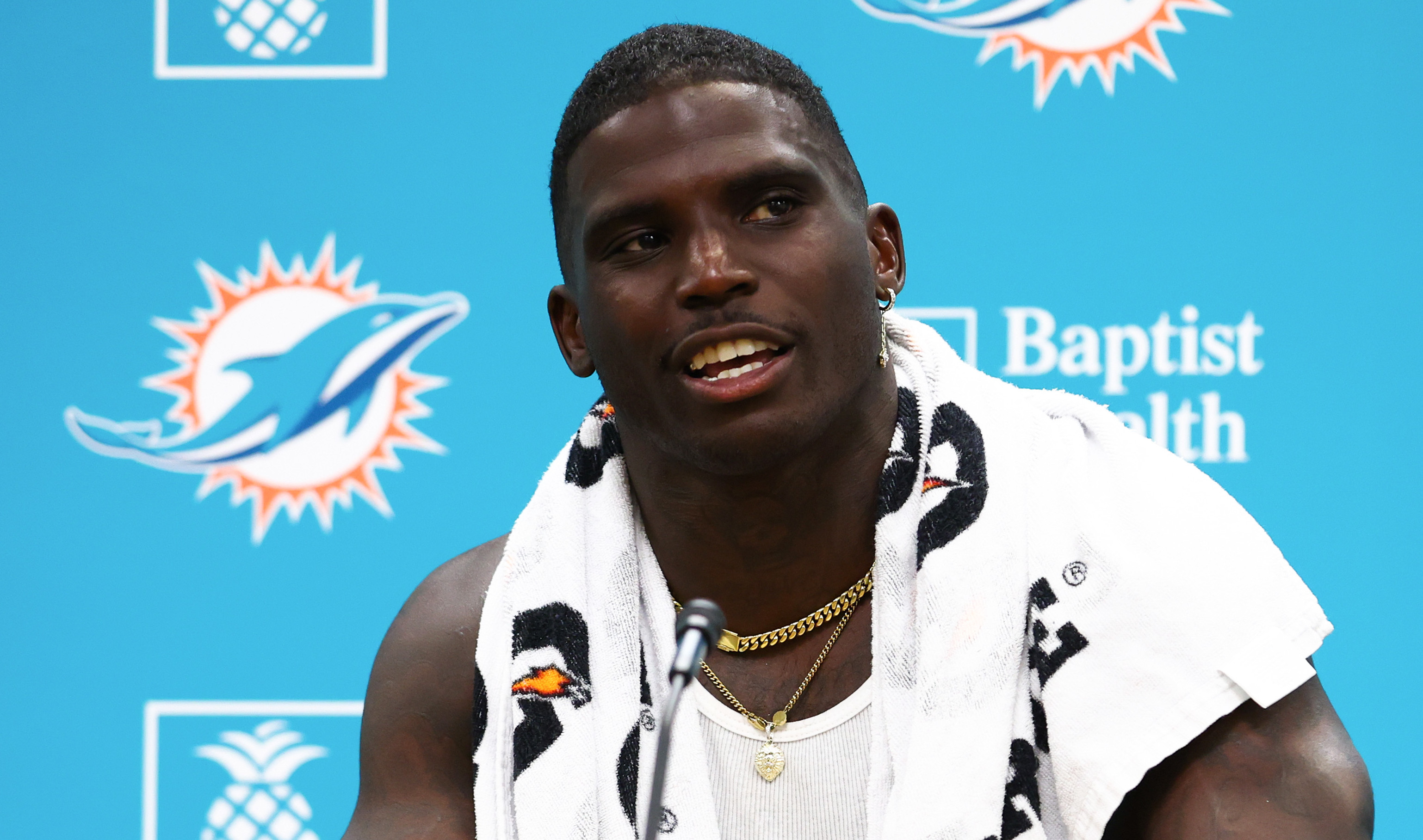 Tyreek Hill at a press conference, wearing a tank top and towel, with a Miami Dolphins backdrop.