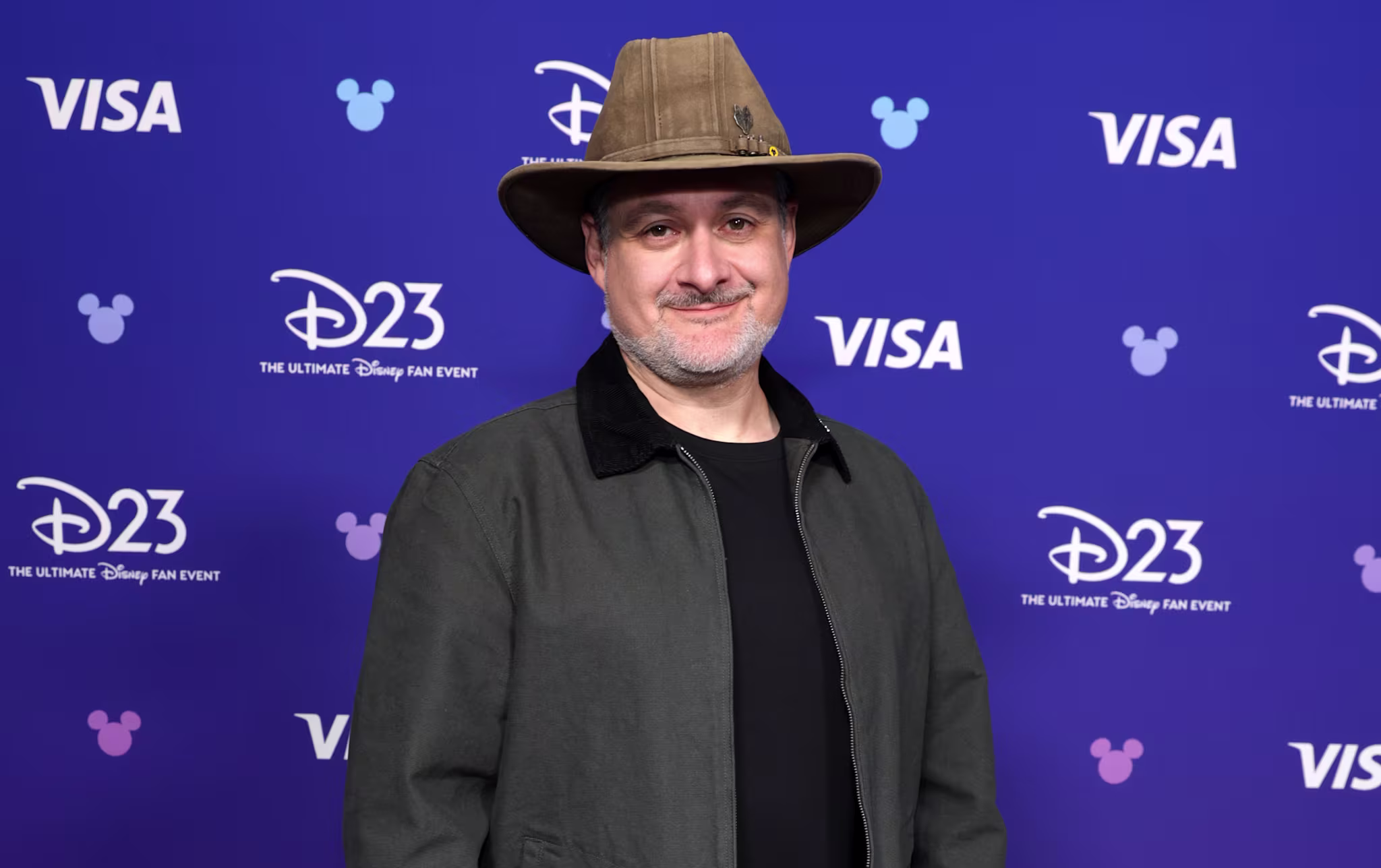 Dave Filoni, in a hat and casual attire, stands on a blue carpet at a D23 event backdrop.