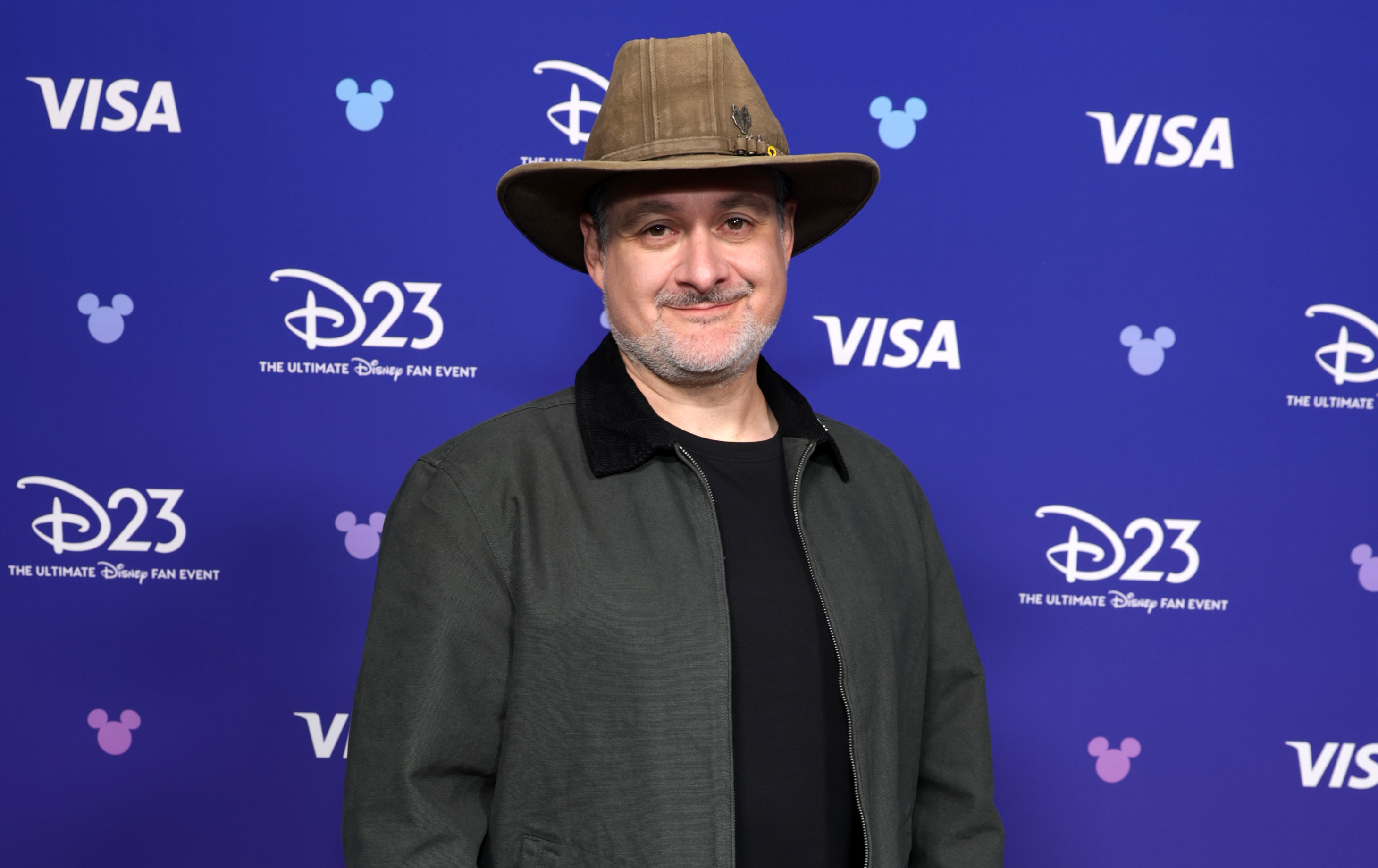 Dave Filoni, in a hat and casual attire, stands on a blue carpet at a D23 event backdrop.