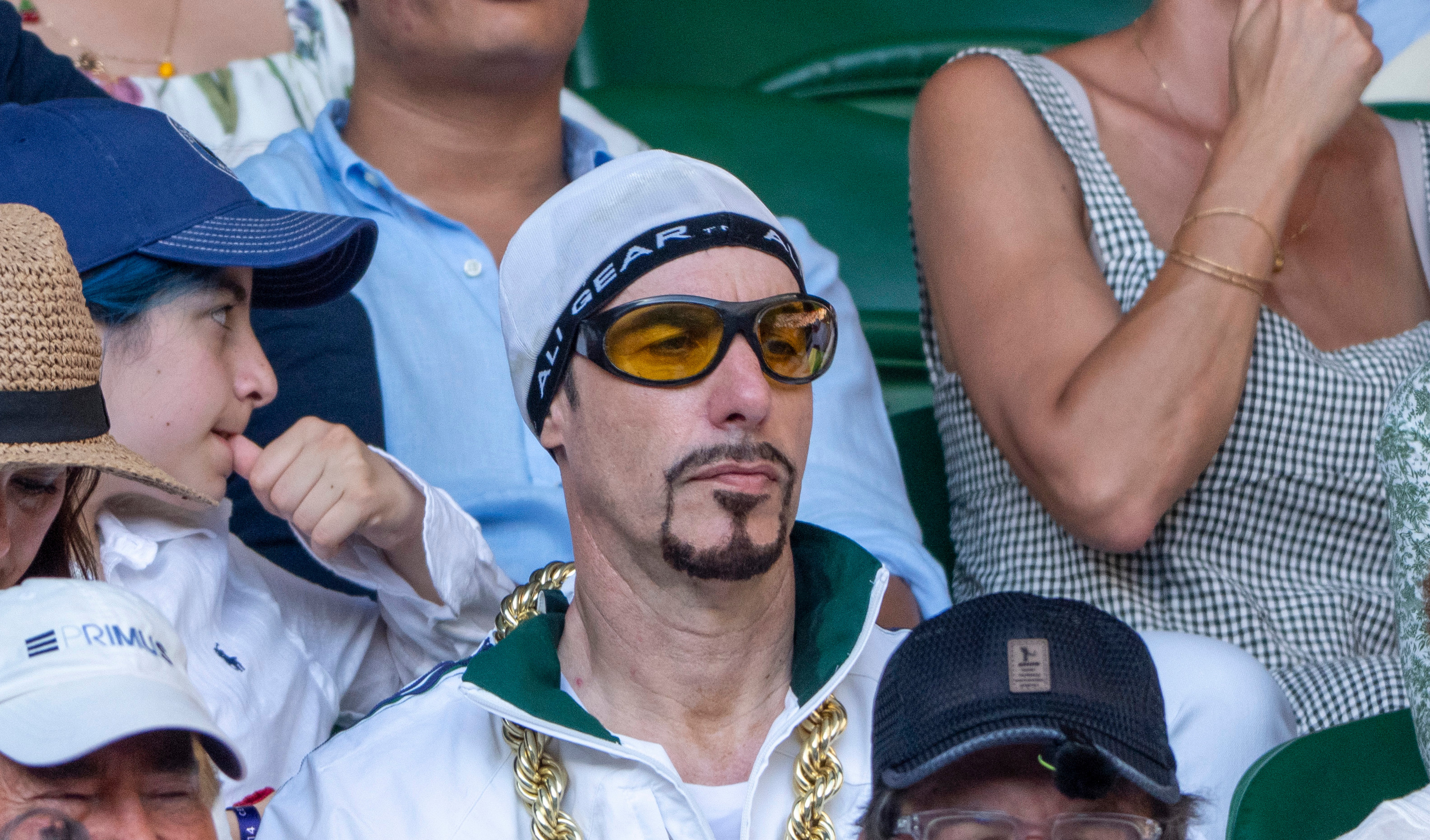 A man dressed as Ali G, wearing a white hoodie, gold chain, and sunglasses, sits among a crowd at an outdoor event.