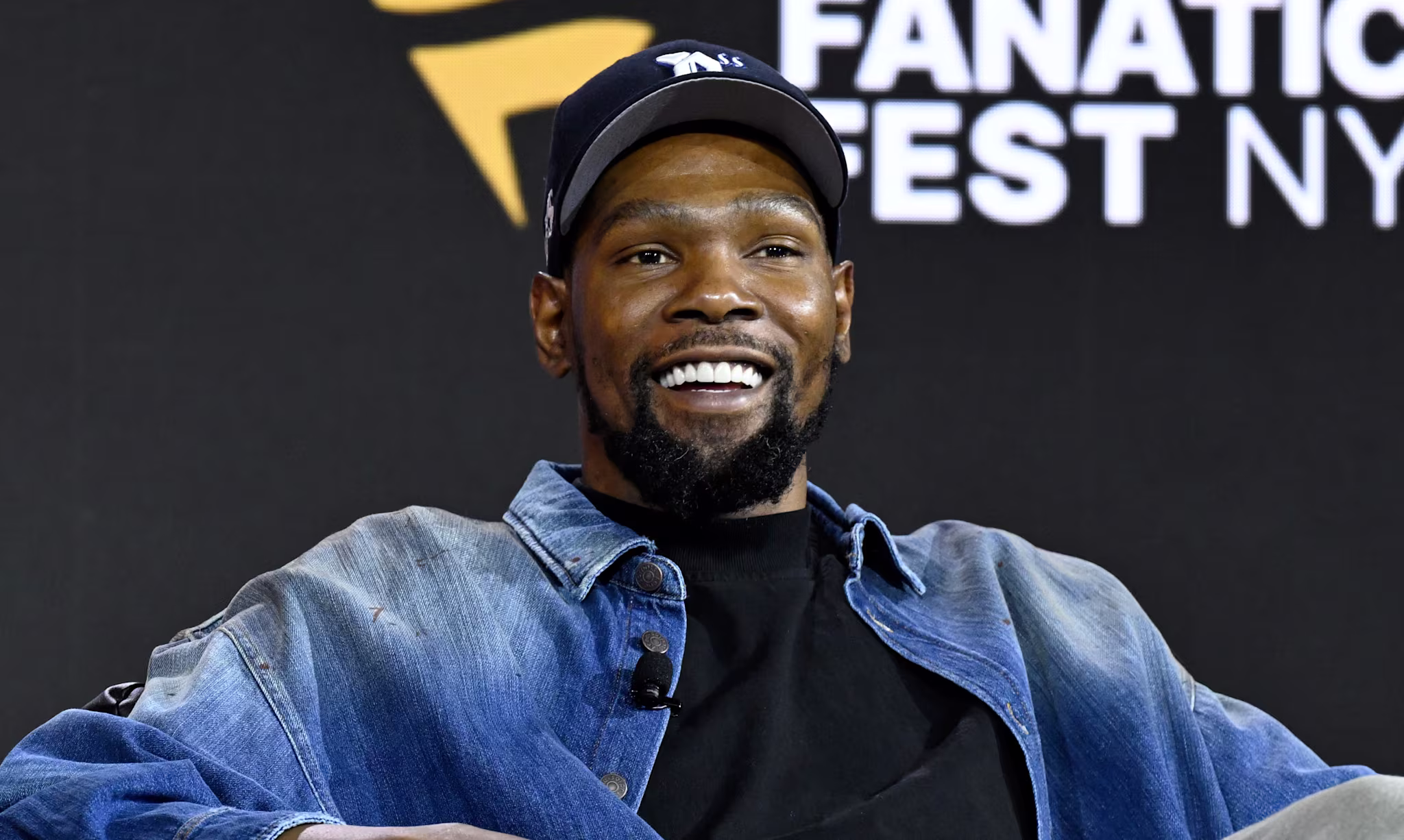 Kevin Durant, wearing a denim jacket and cap, smiles while seated at Fanatics Fest NYC.