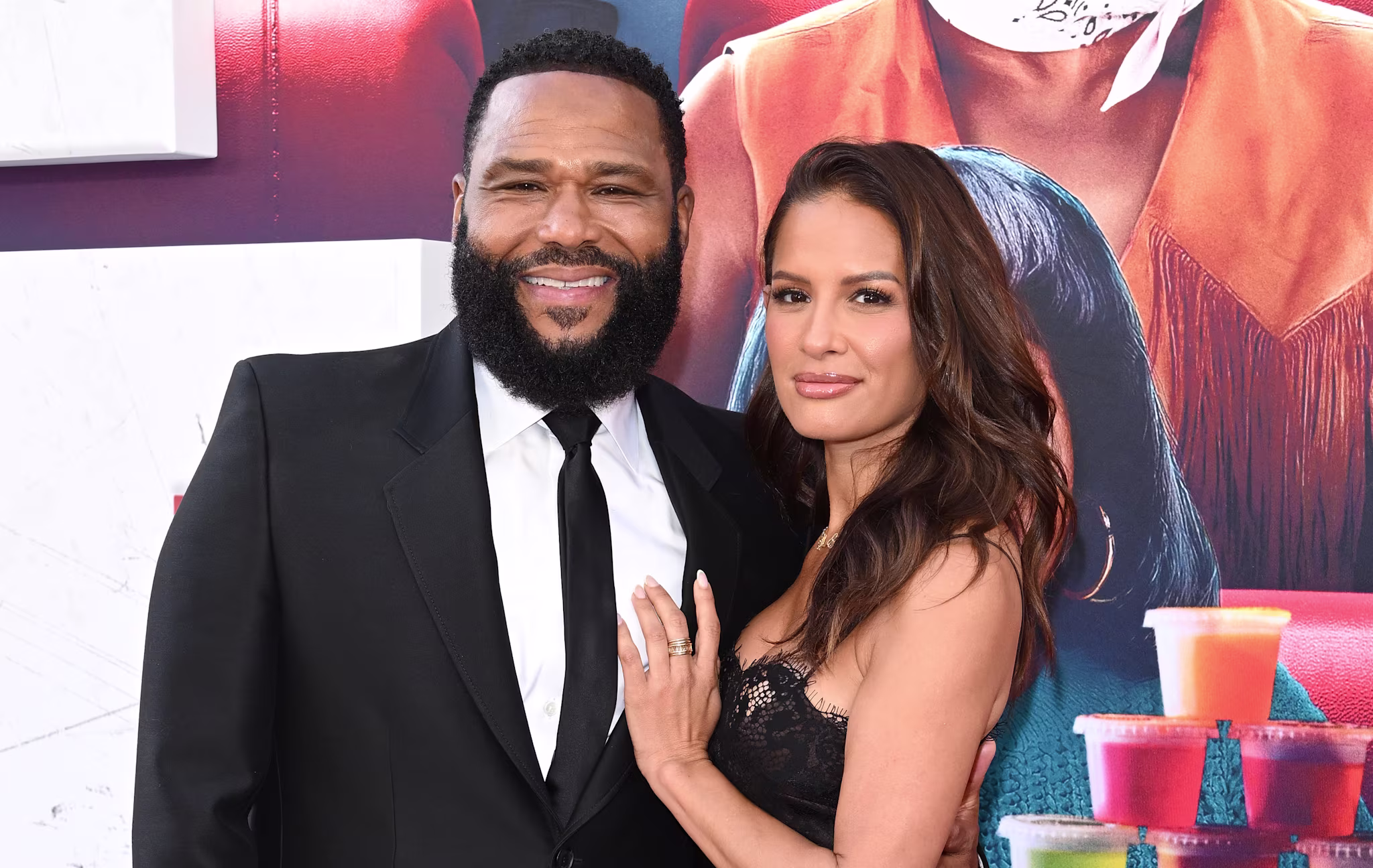 Anthony Anderson in a black suit and Rocsi Diaz in a black lace dress pose together on a red carpet, smiling.