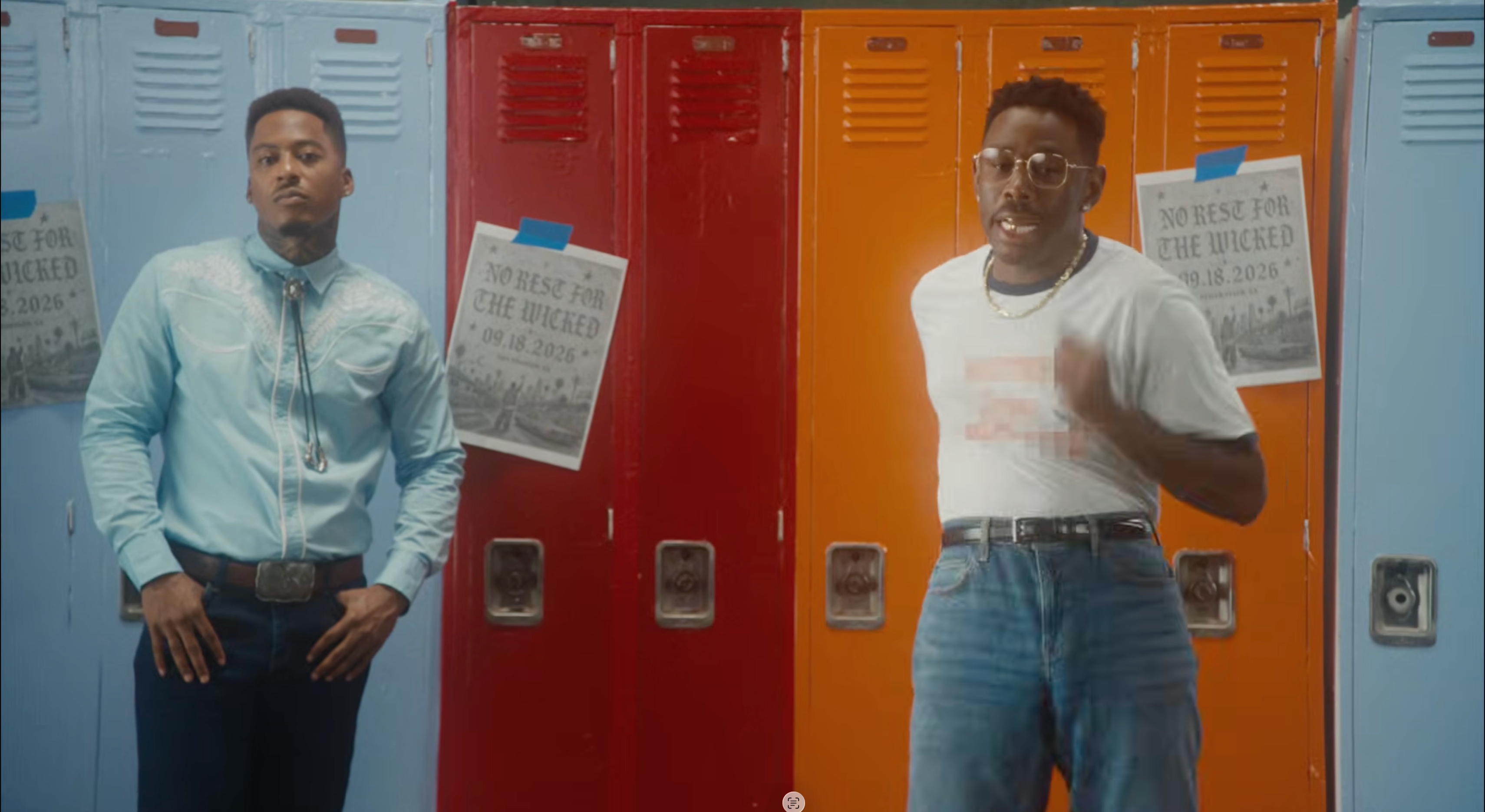 AZ Chike and Tyler, the Creator stand in front of colorful lockers. Chike wears a blue shirt; Tyler wears a white t-shirt and glasses. Posters are on the lockers.