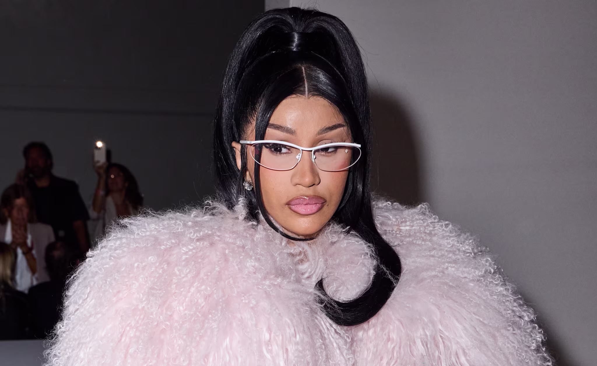 Cardi B in a fluffy pink coat and glasses, with a high ponytail, attending an event.
