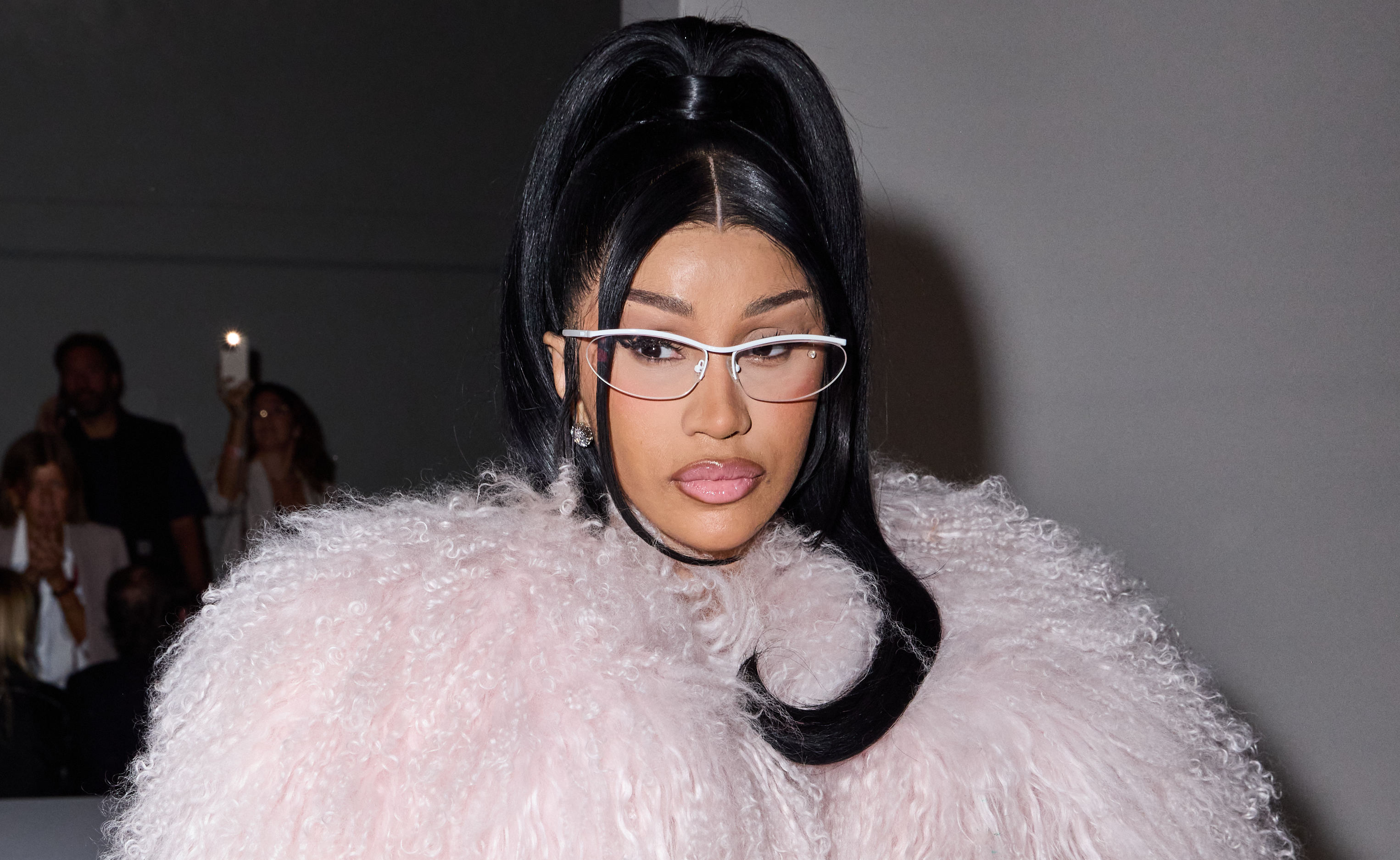 Cardi B in a fluffy pink outfit, wearing glasses, with long black hair styled in a high ponytail.