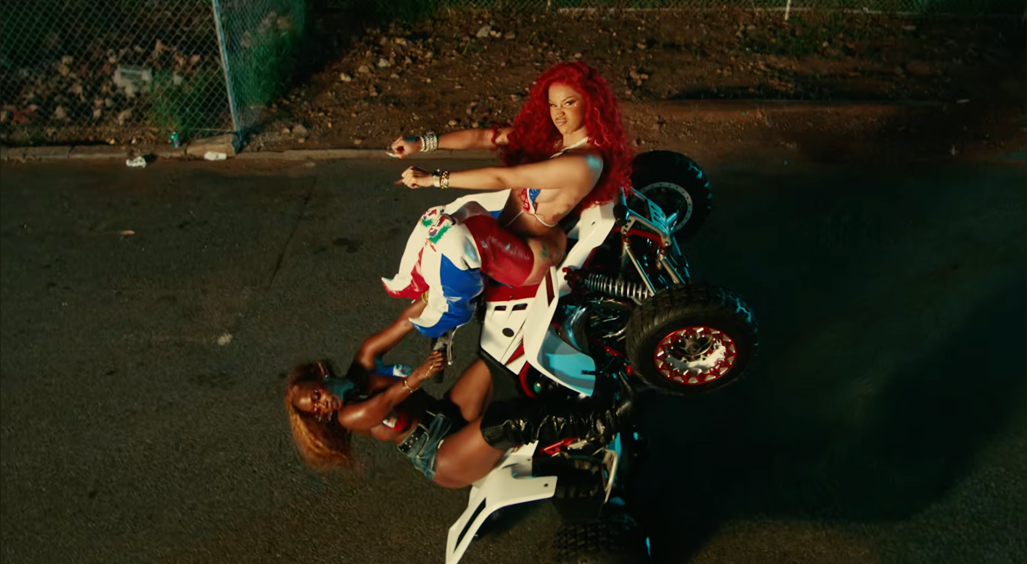 Two women on an ATV, one with bright red hair and colorful outfit, the other with dark hair and denim shorts, in an urban setting.
