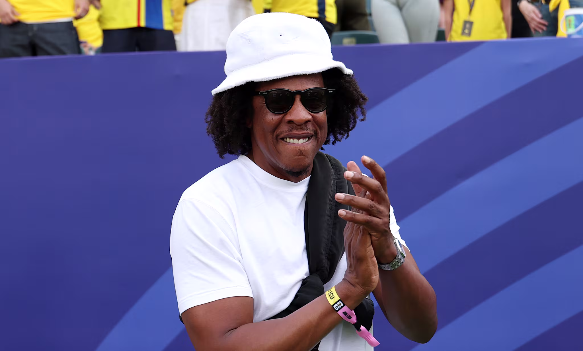 Jay-Z in a white hat and sunglasses claps, standing in front of a crowd wearing yellow jerseys at a sports event.