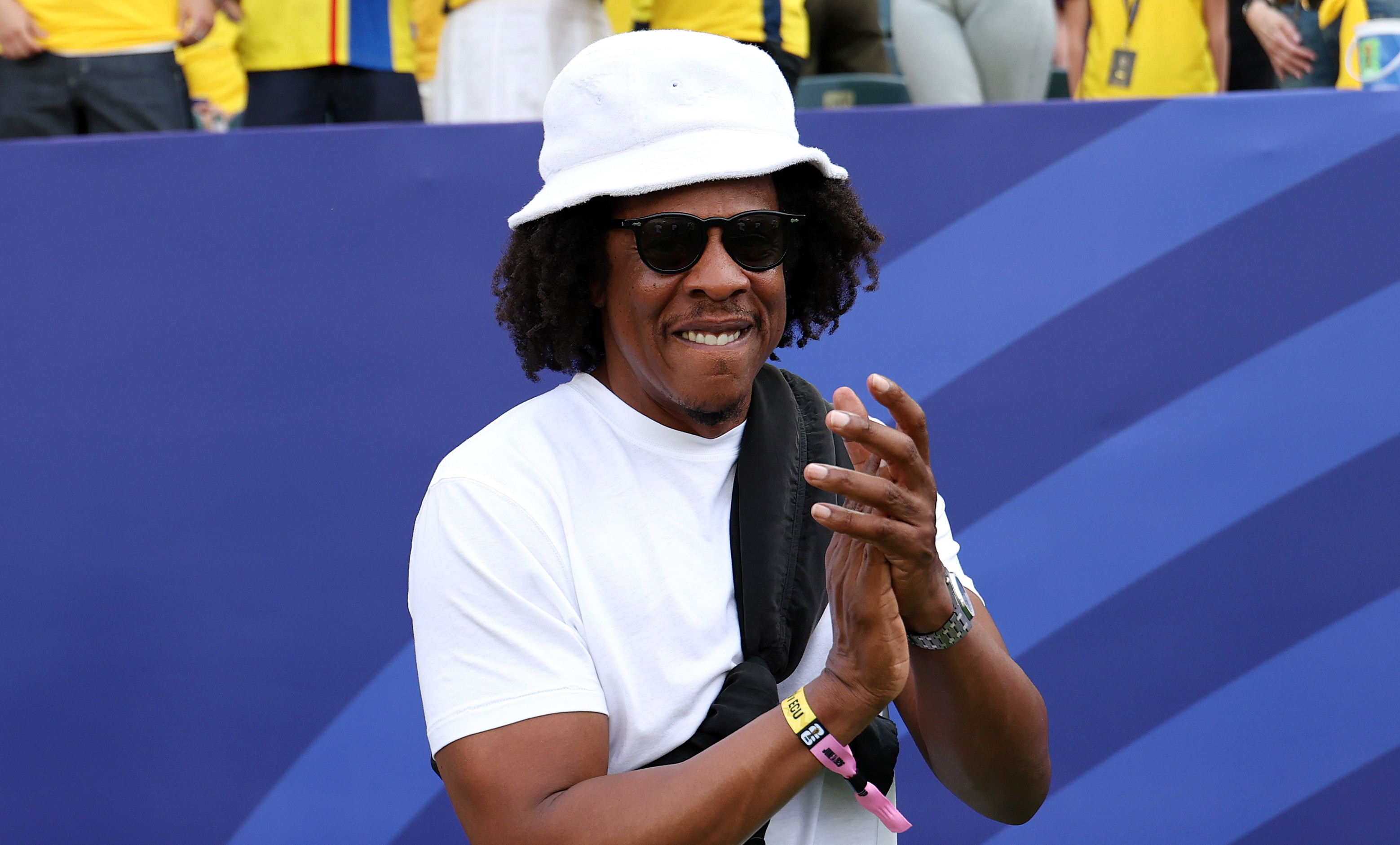 Jay-Z in a white hat and sunglasses claps, standing in front of a crowd wearing yellow jerseys at a sports event.