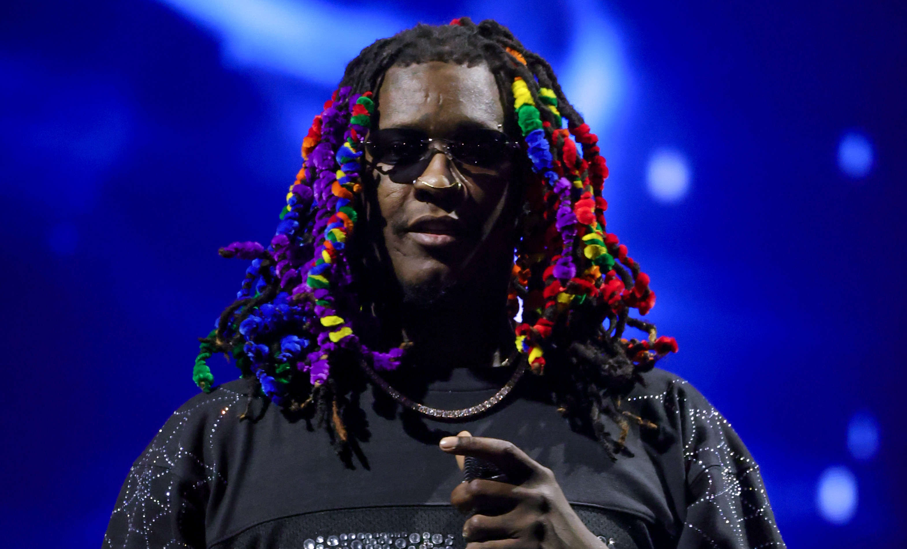 Young Thug with colorful hair and sunglasses on stage, wearing a black outfit adorned with sparkles.