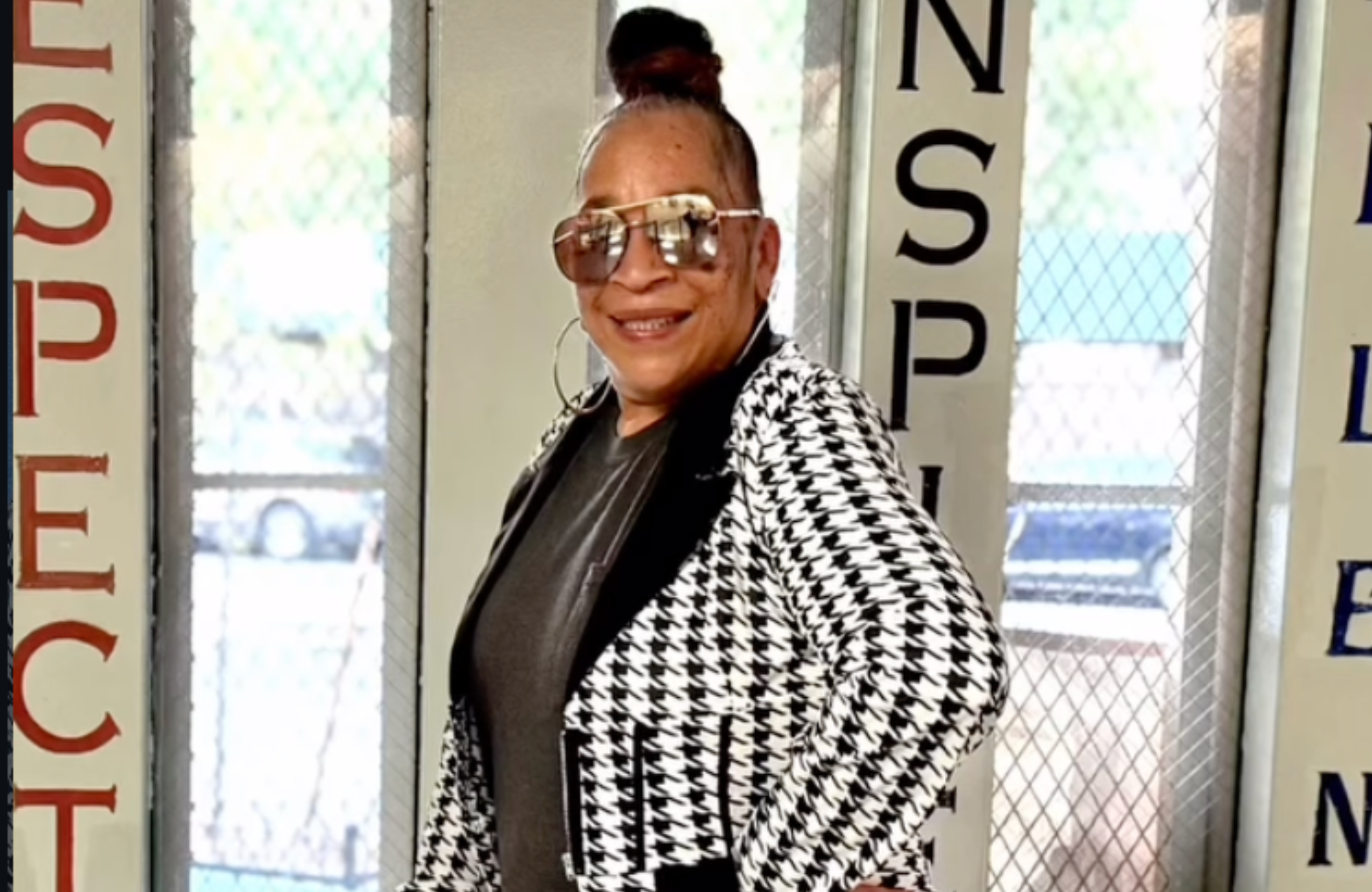 MC Sparky D wearing sunglasses and a houndstooth jacket smiles, standing in front of a wall with motivational words.