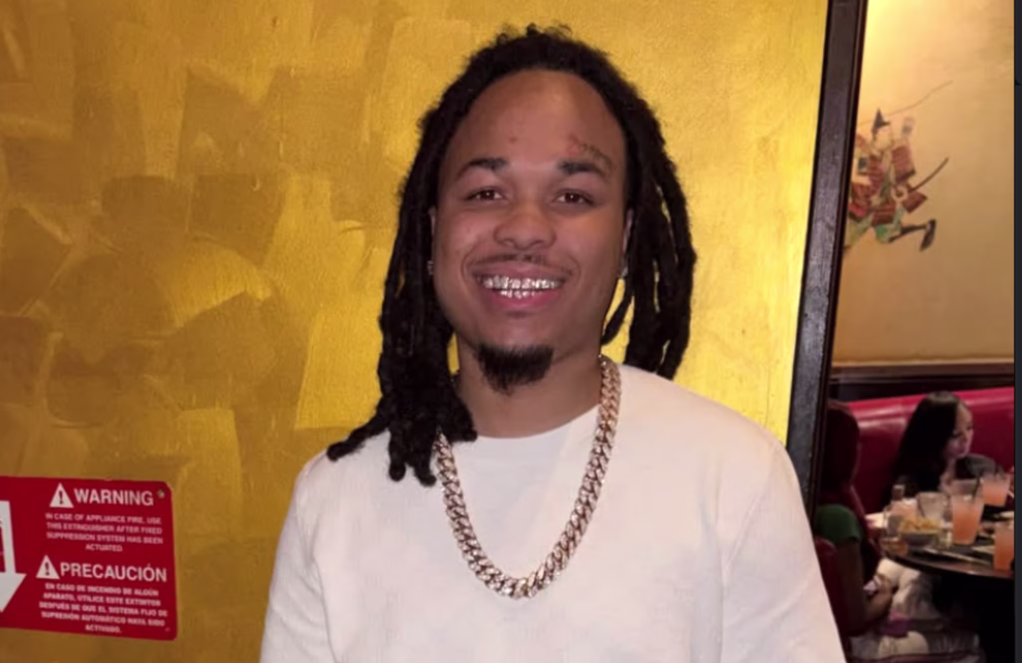 Bloodhound Q50 with dreadlocks and a gold chain smiles in front of a yellow wall, with a restaurant scene in the background.