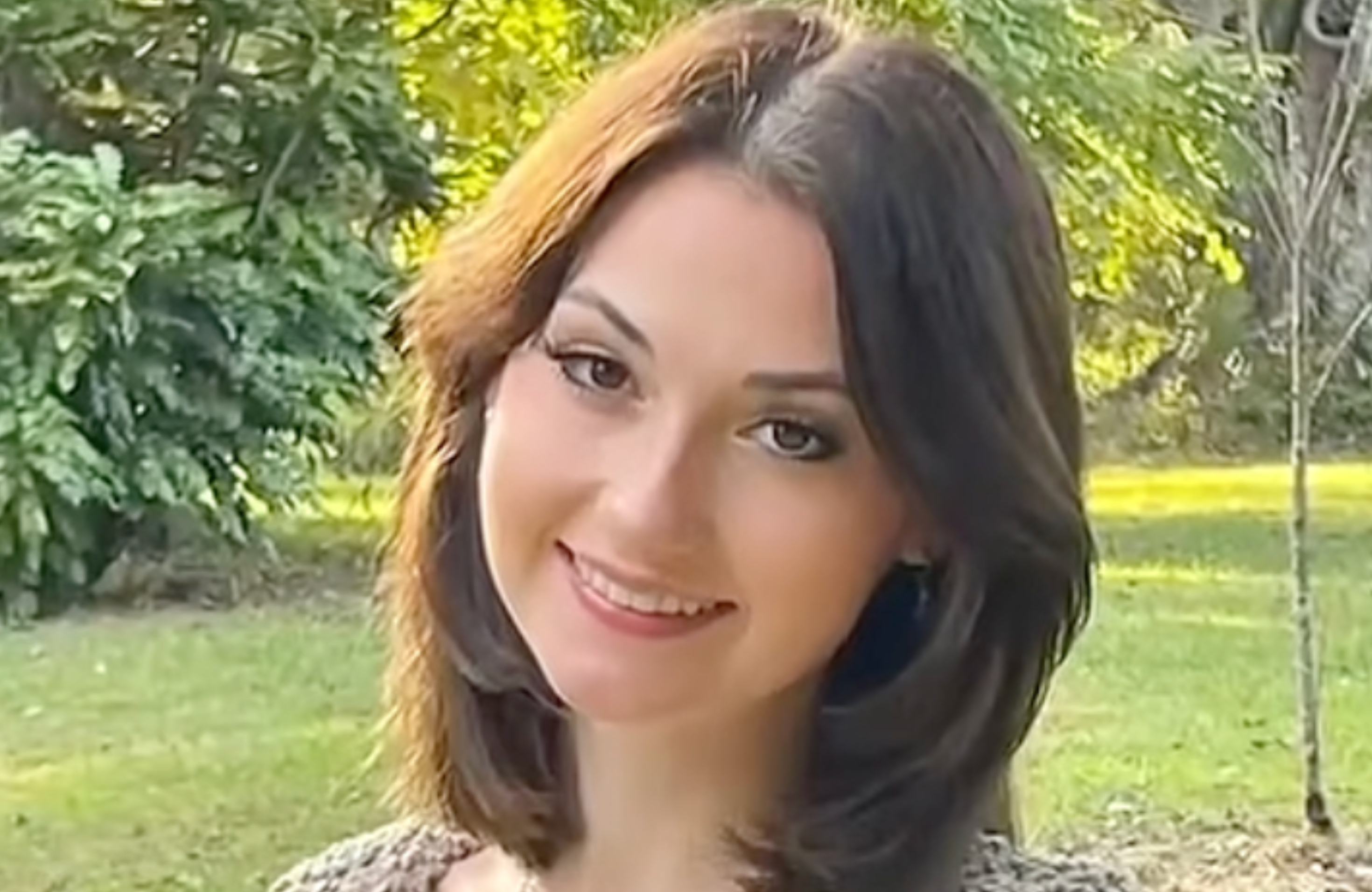 A woman with shoulder-length brown hair smiling in a green park setting.