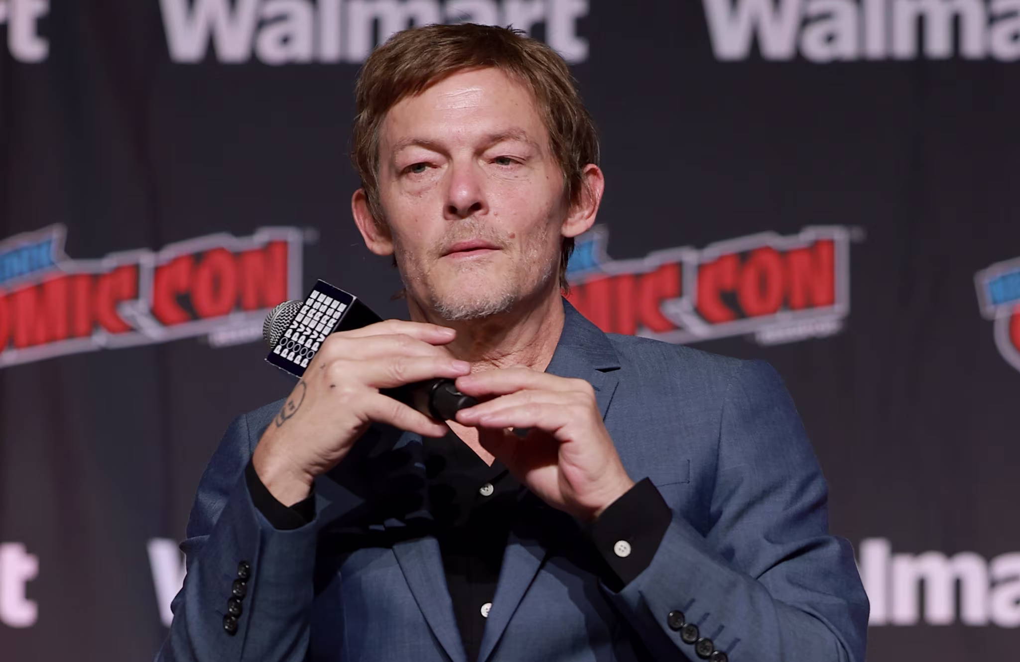 Norman Reedus in a blue suit sits on a director's chair at a Comic Con event, holding a microphone.