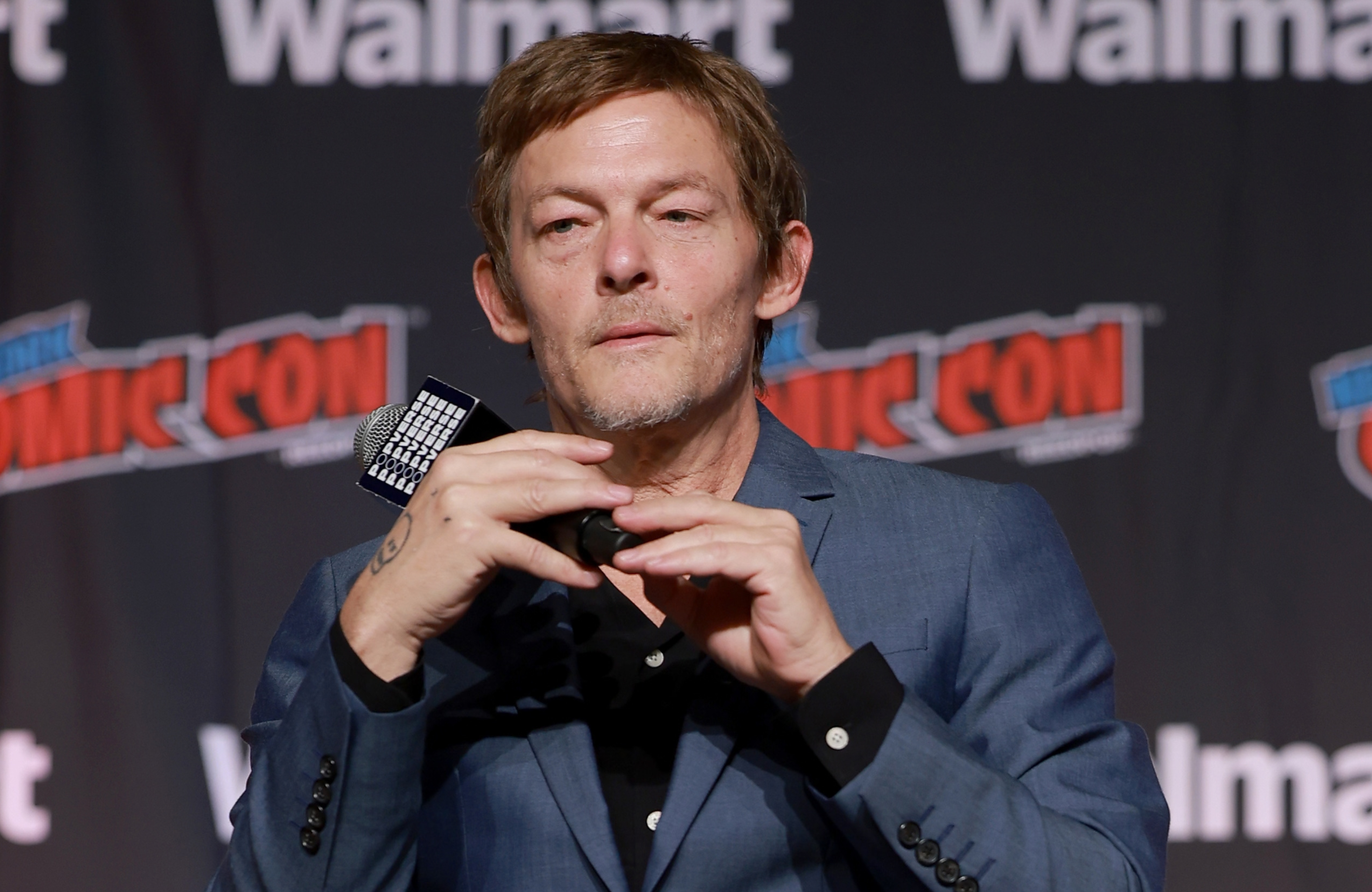 Norman Reedus in a blue suit sits on a director's chair at a Comic Con event, holding a microphone.