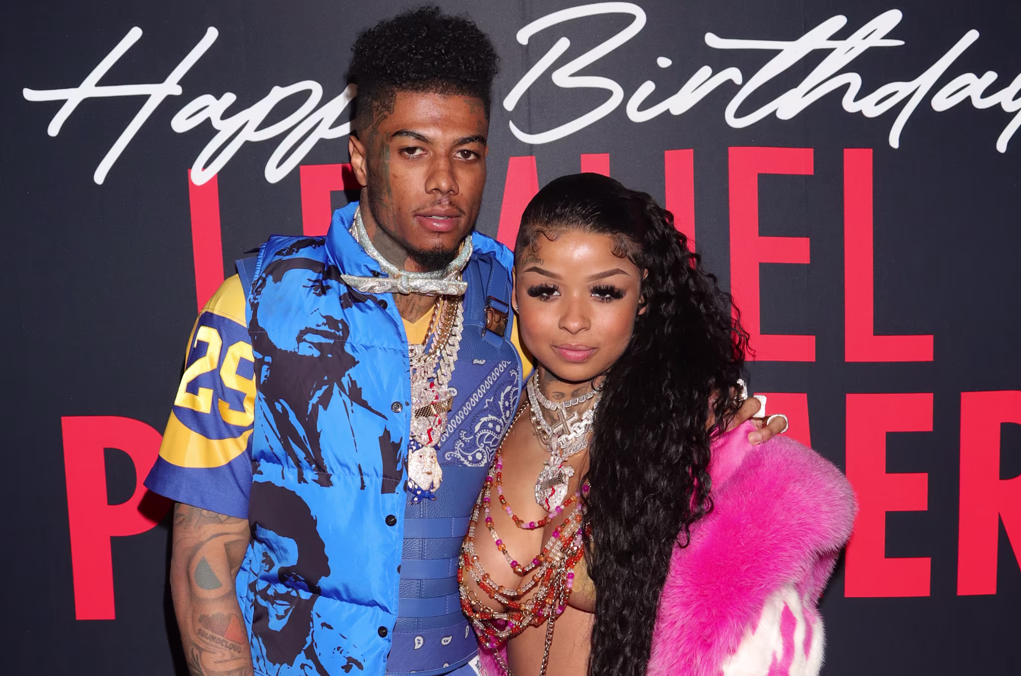 Blueface and Chrisean Rock pose together at an event. The man wears a blue patterned outfit, and the woman wears a beaded top with a pink fur coat.