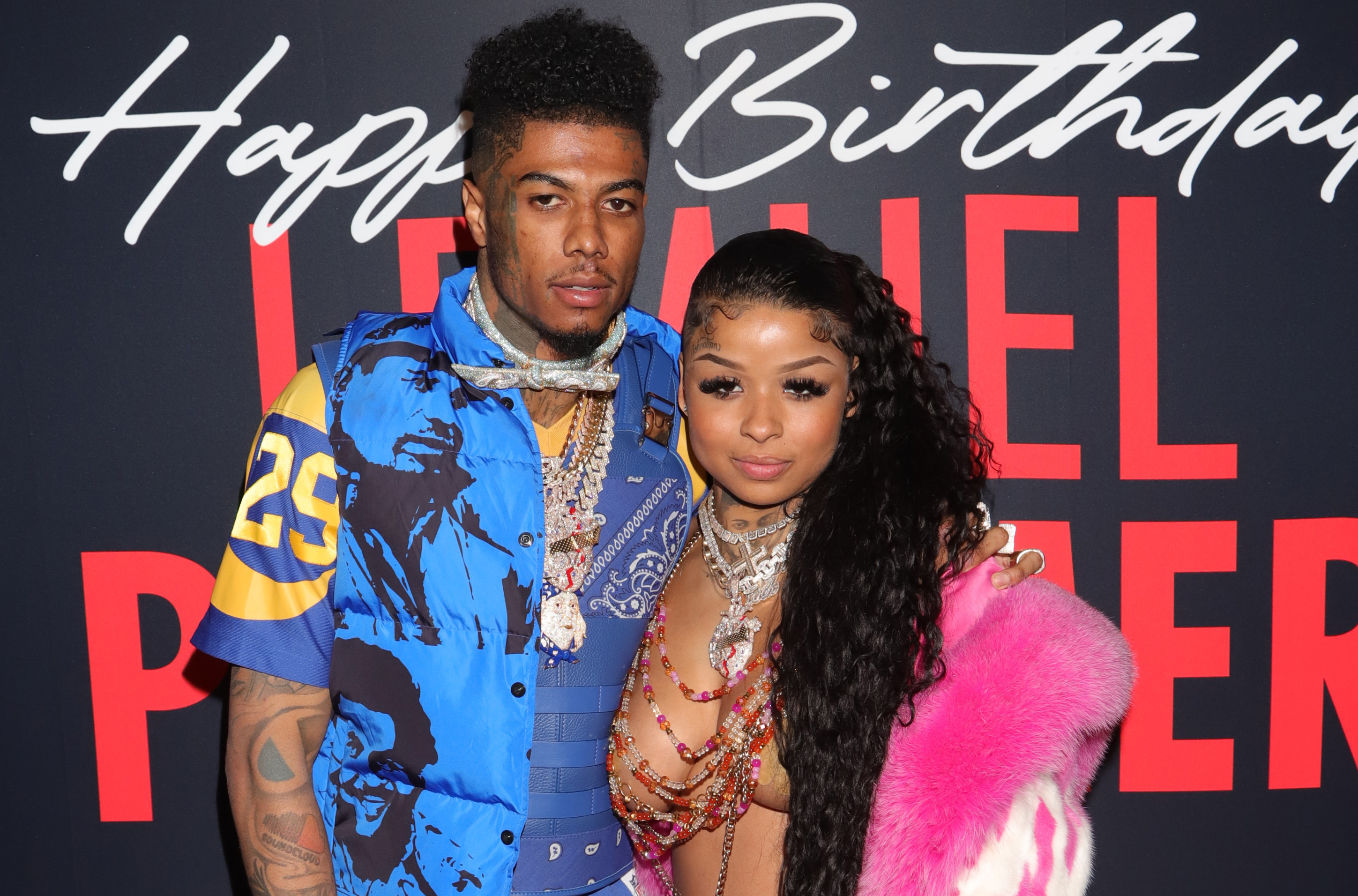 Blueface and Chrisean Rock pose together at an event. The man wears a blue patterned outfit, and the woman wears a beaded top with a pink fur coat.