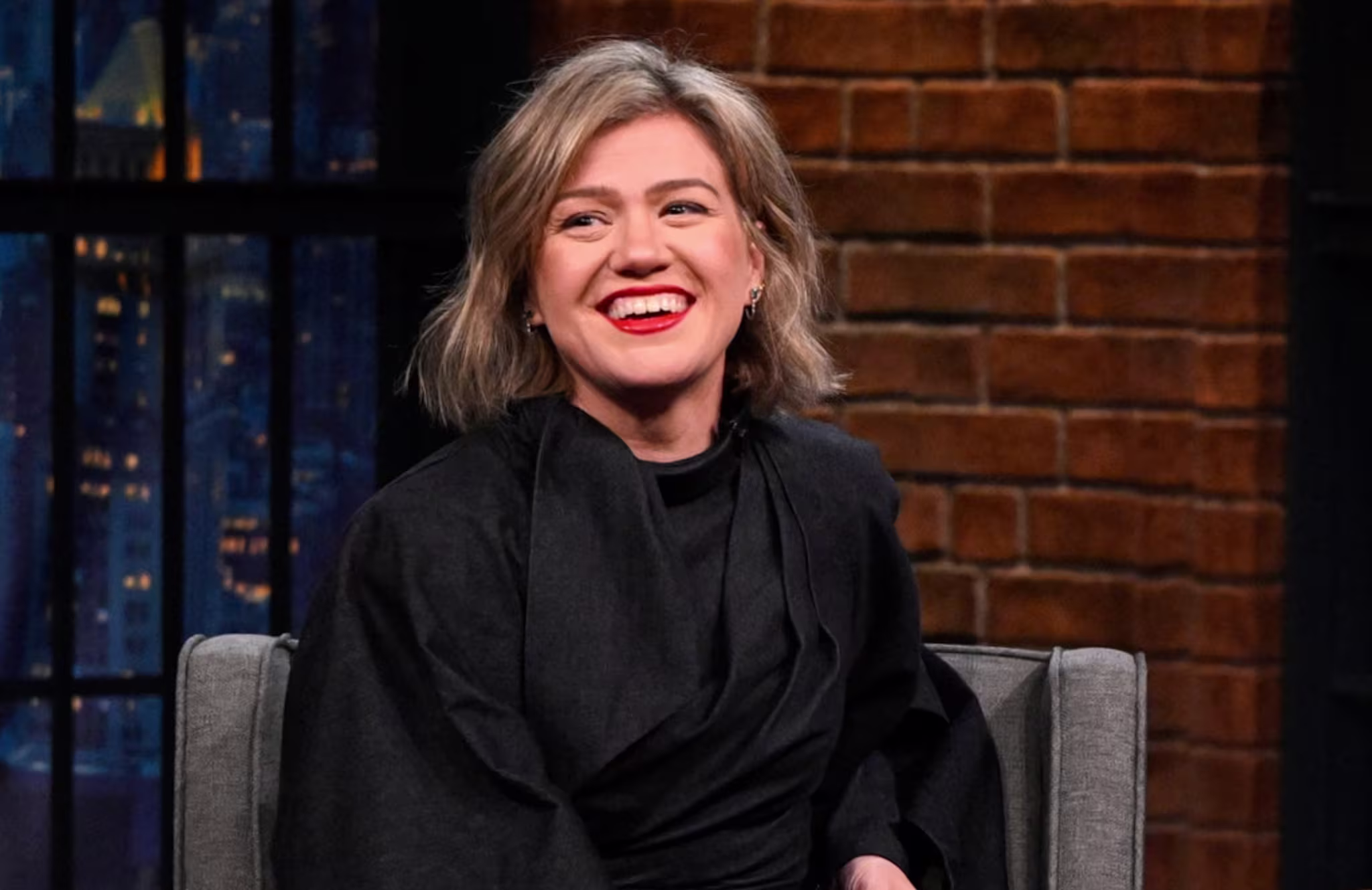 Kelly Clarkson smiling, seated on a talk show set, wearing a black outfit. Brick wall and window in the background.