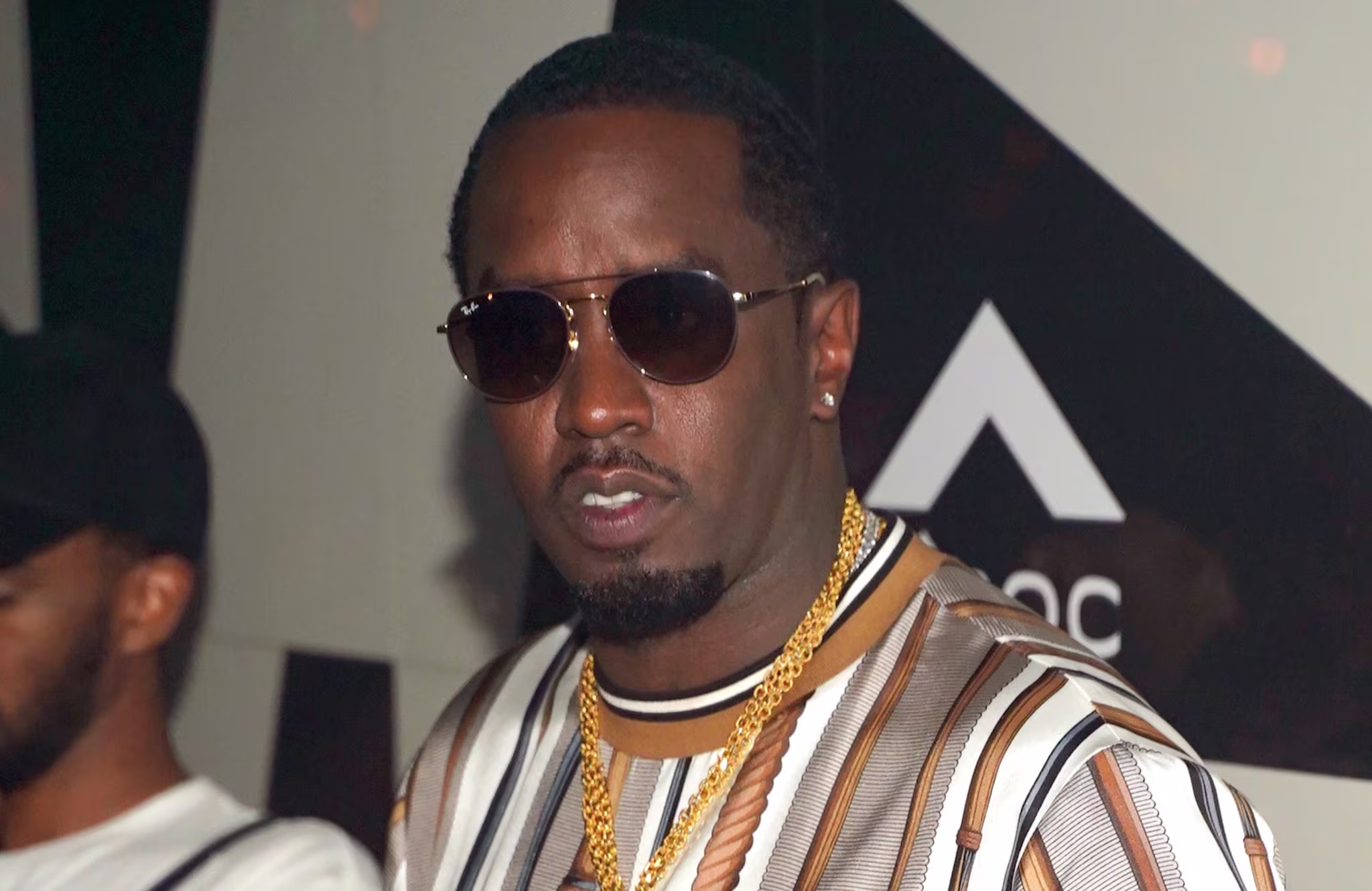 Diddy wearing sunglasses, a striped shirt, and gold chains at an event.