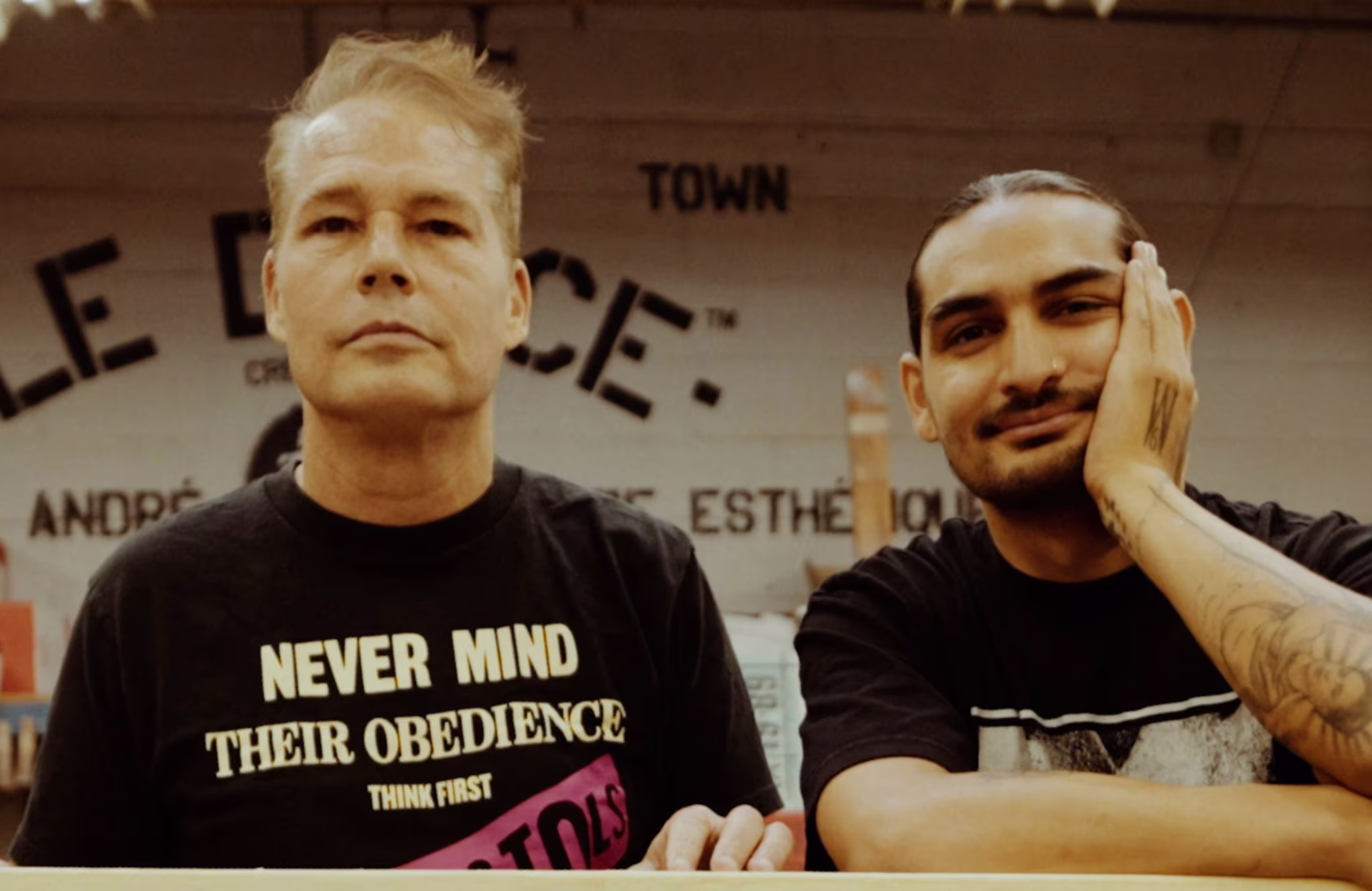 Two people are standing indoors. One wears a "Never Mind Their Obedience" shirt, and the other rests his face on his hand, showing a tattoo.