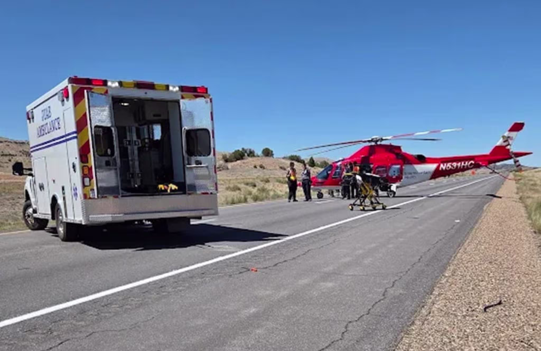 An ambulance and a red helicopter are on a road. Emergency responders are nearby, with a stretcher visible. Clear blue sky.