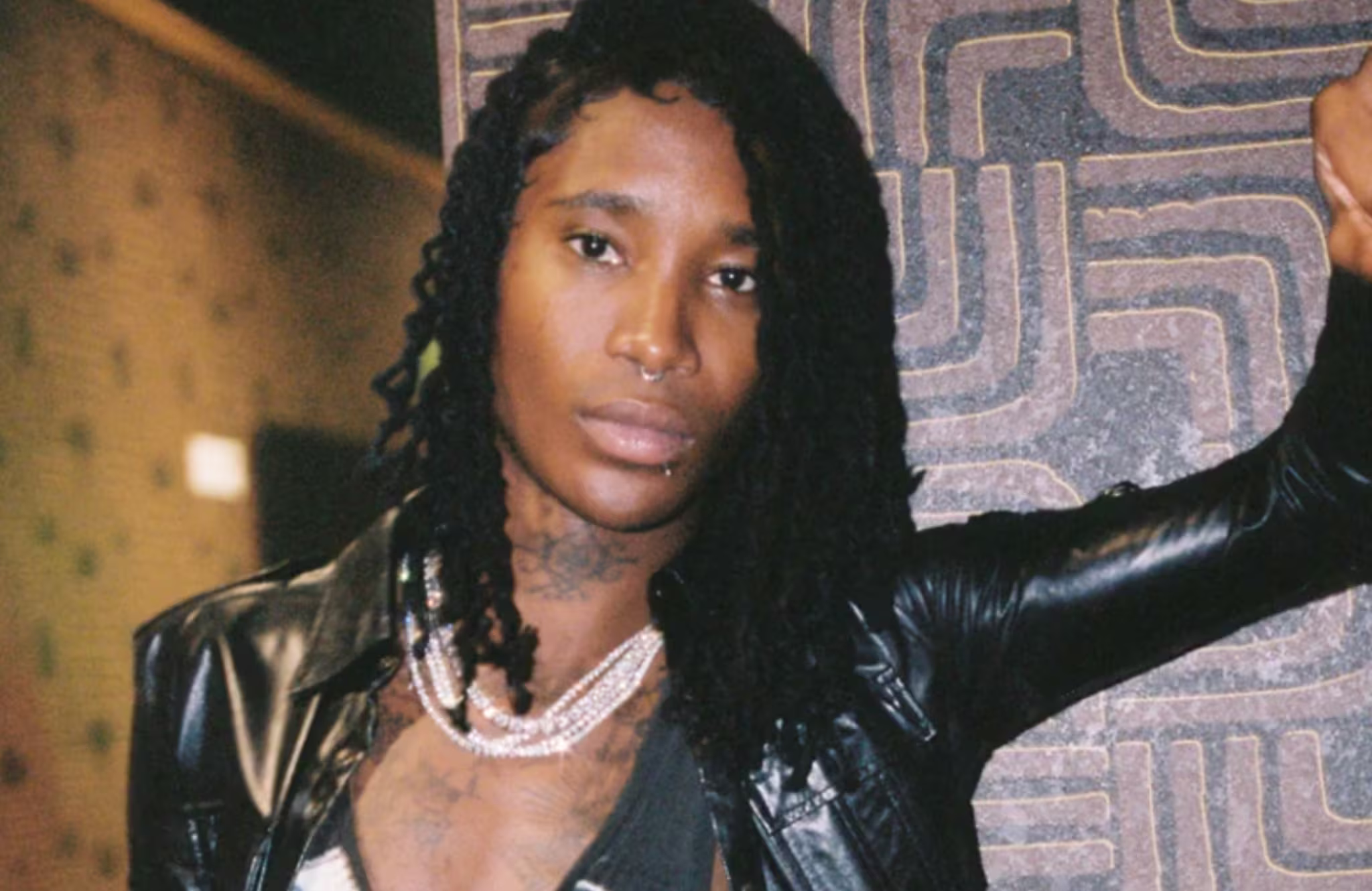 Nine Vicious with long braided hair, wearing a leather jacket and layered necklaces, standing against a patterned wall.