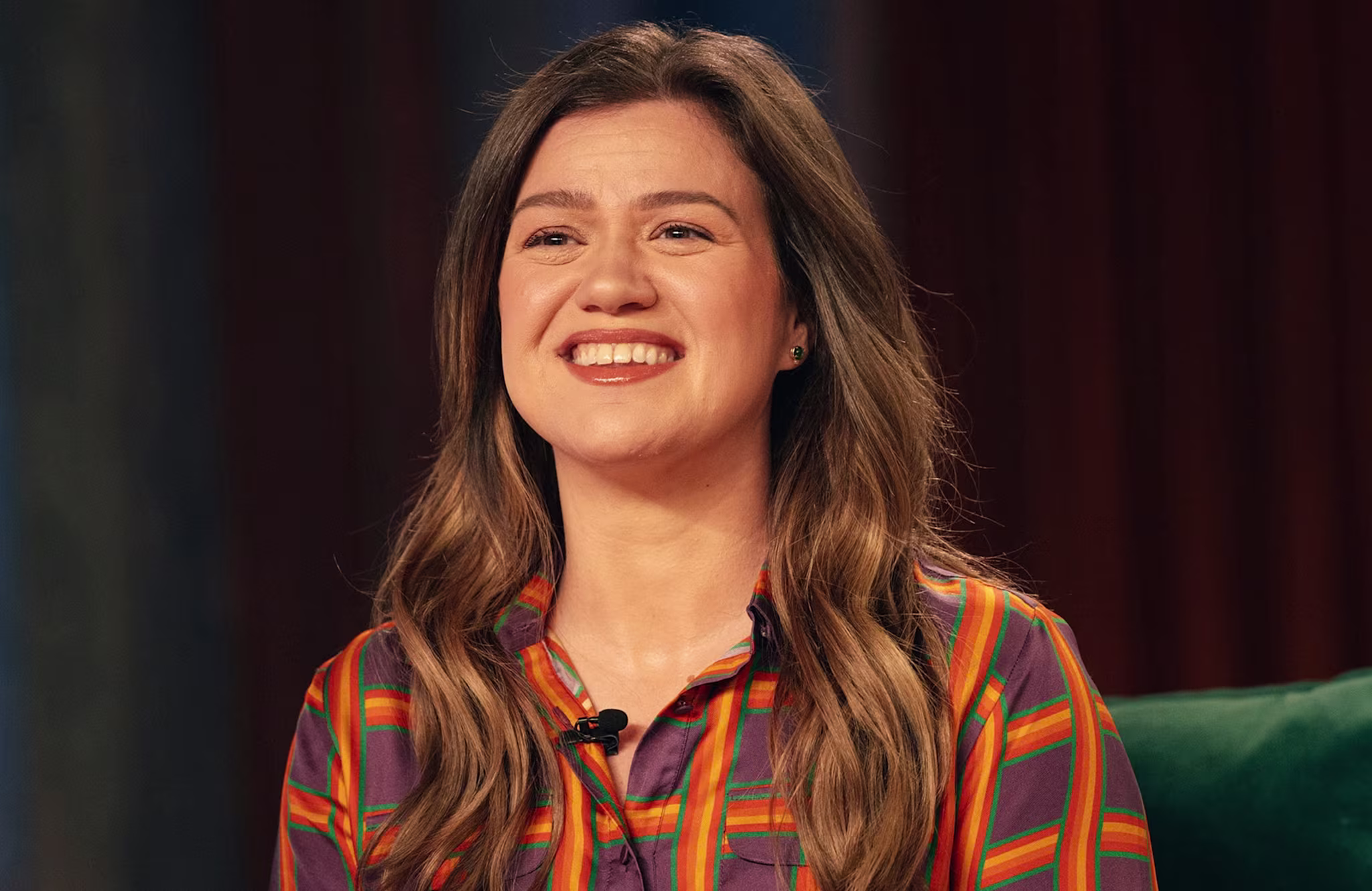 Kelly Clarkson with long brown hair, wearing a colorful striped shirt, smiling.