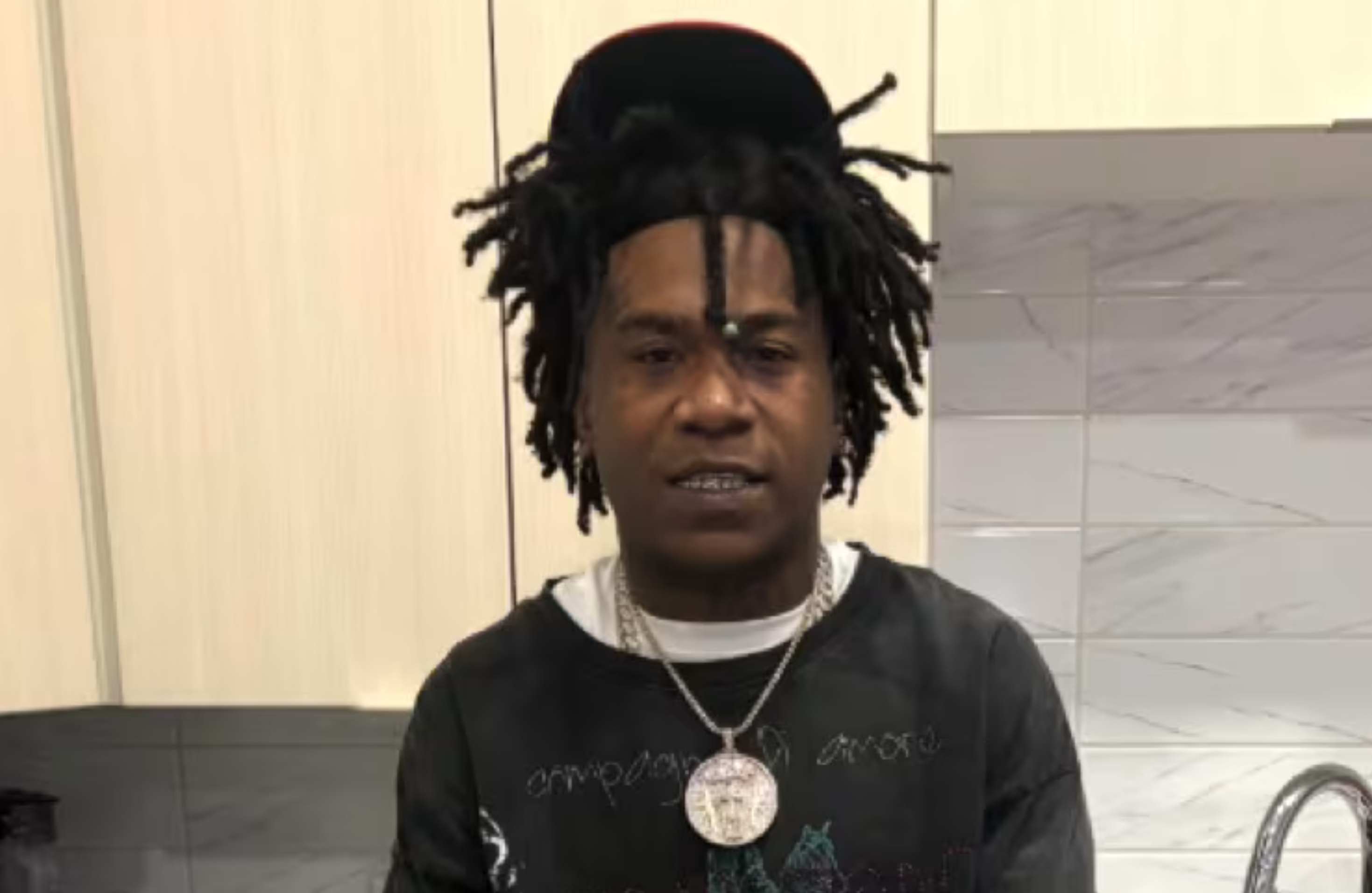 NBA Ben10 with dreadlocks and a cap, wearing a necklace, stands in a kitchen.