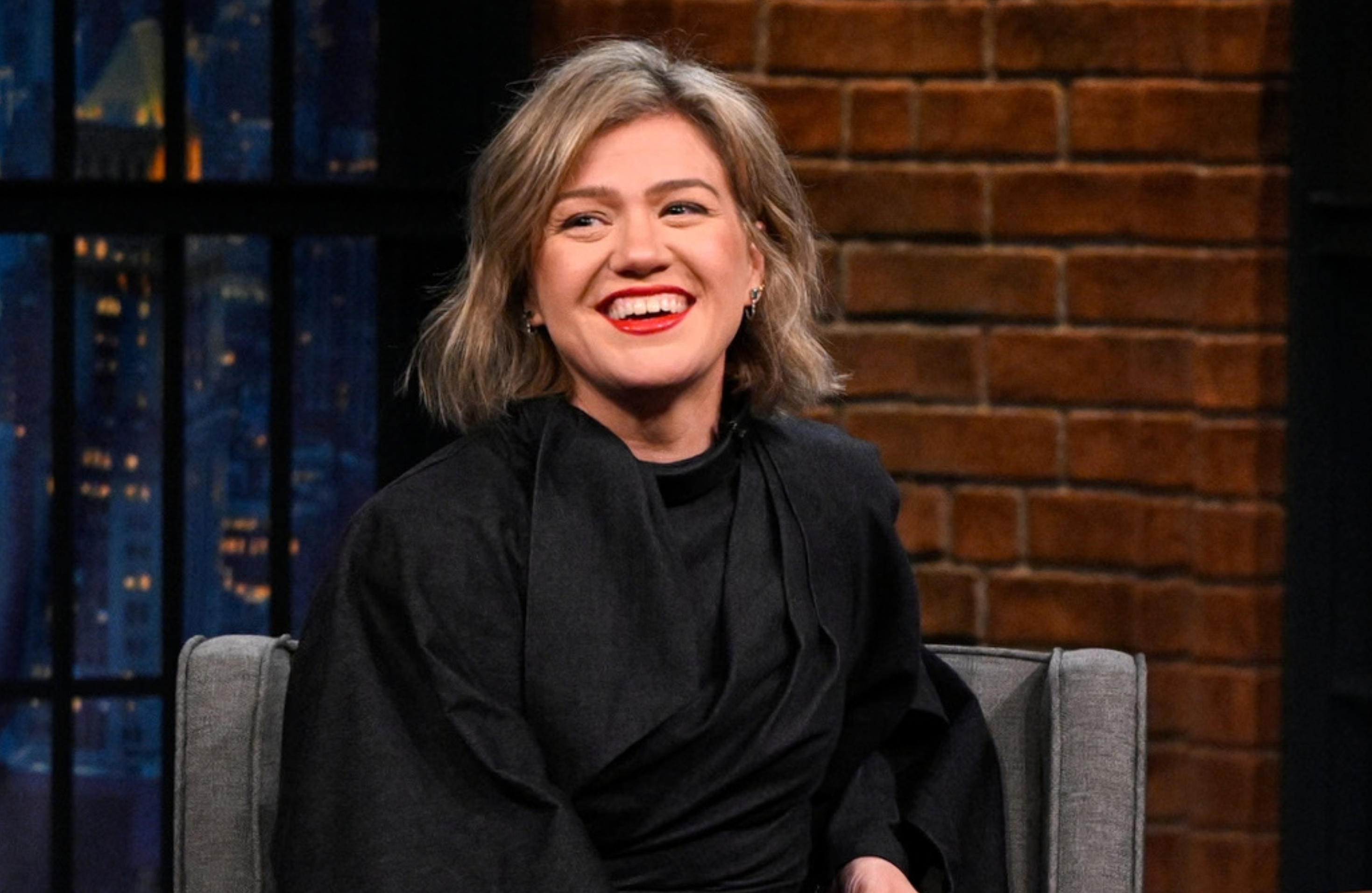 Kelly Clarkson smiling, seated on a talk show set, wearing a black outfit. Brick wall and window in the background.