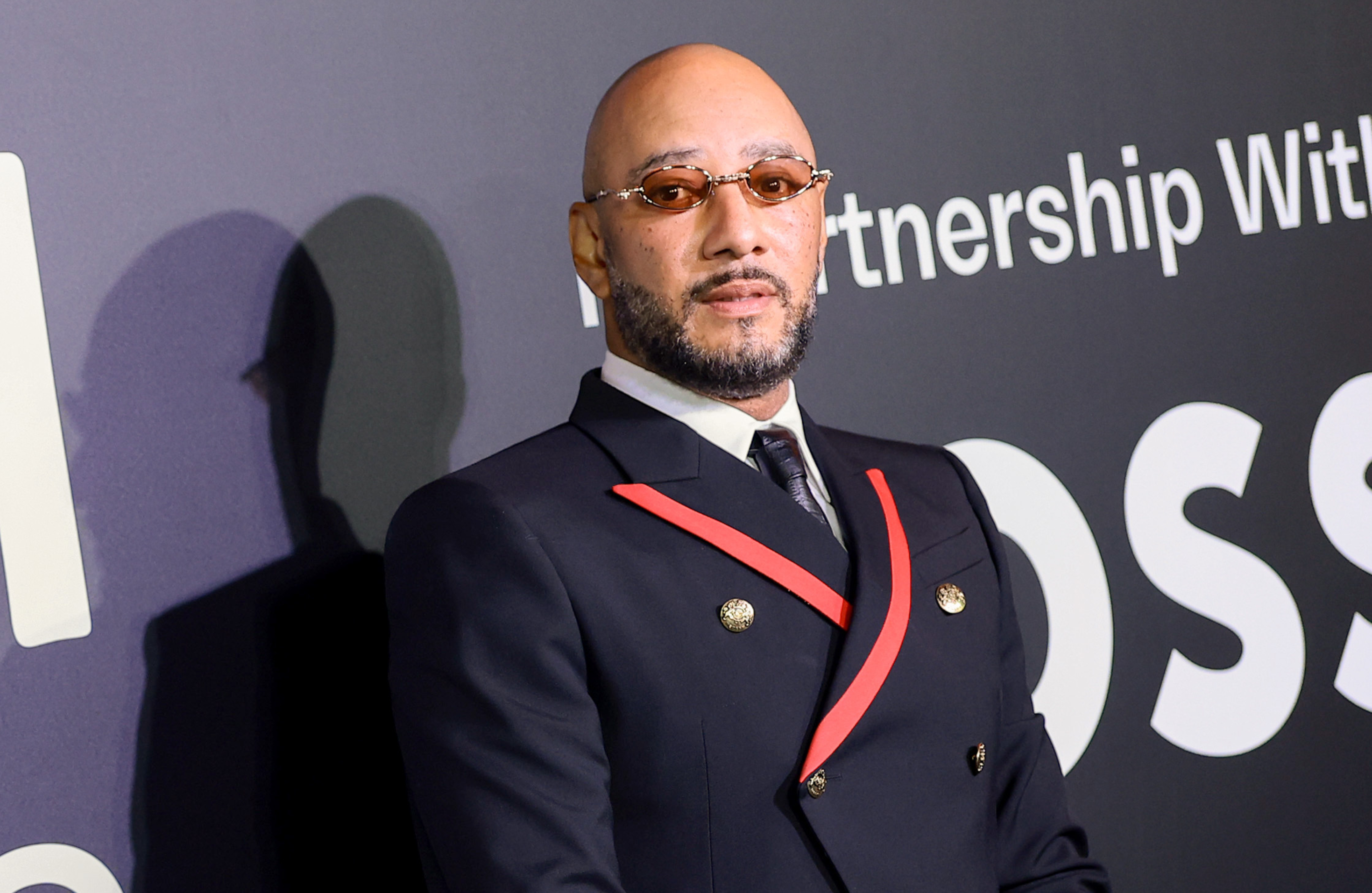 Swizz Beatz in a stylish black suit with red accents and sunglasses at an event, standing against a dark background with text.
