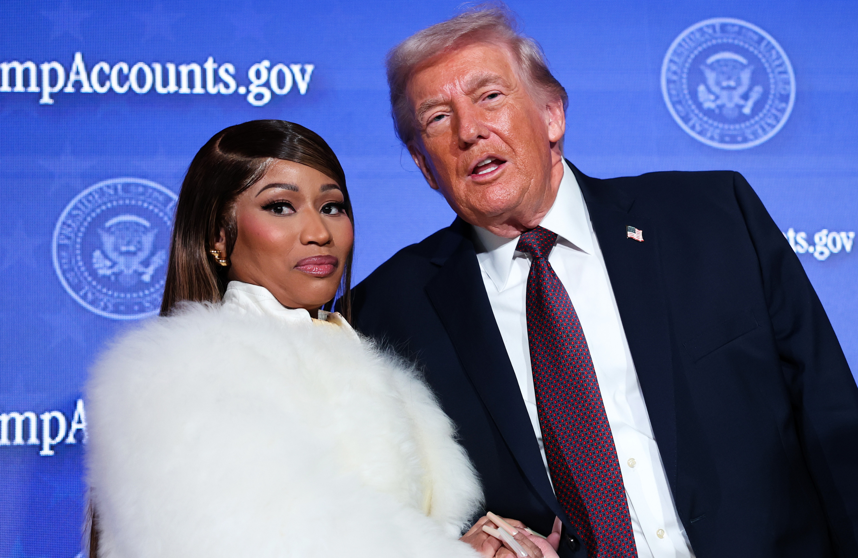 Nicki Minaj in a white fur coat poses with Donald Trump at an event, standing against a blue backdrop.
