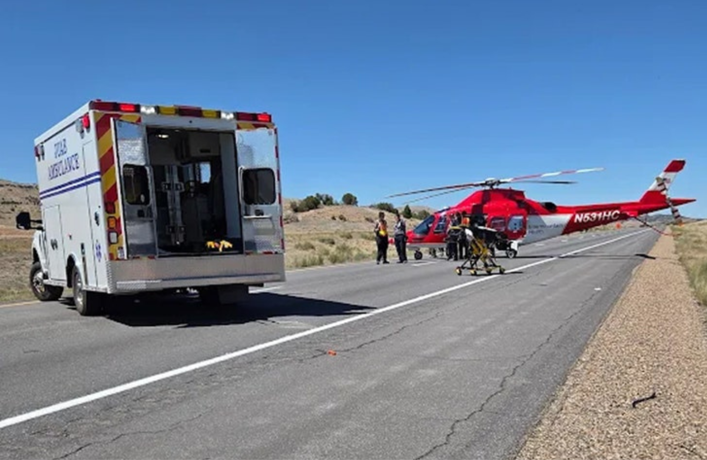 An ambulance and a red helicopter are on a road. Emergency responders are nearby, with a stretcher visible. Clear blue sky.