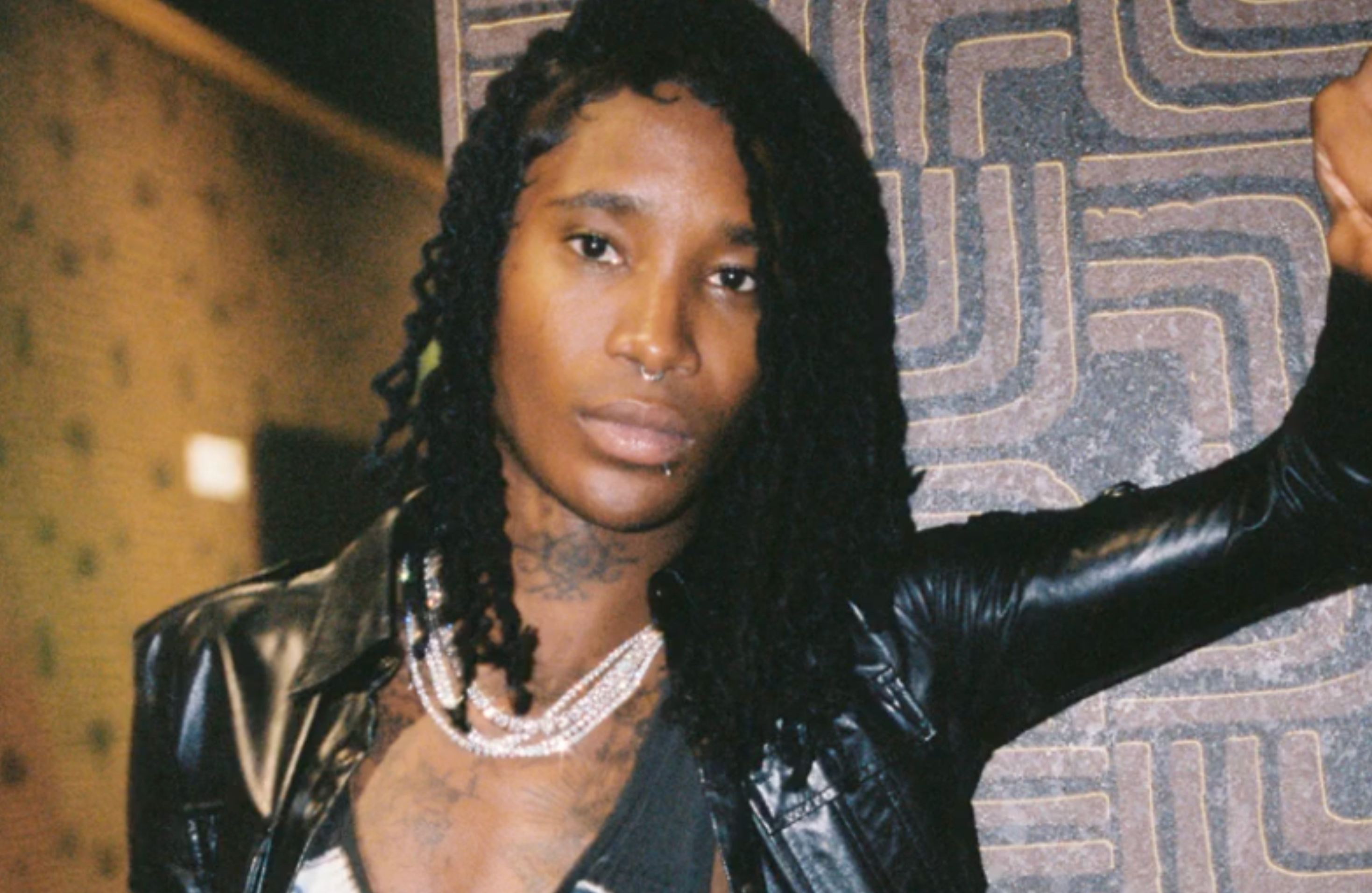 Nine Vicious with long braided hair, wearing a leather jacket and layered necklaces, standing against a patterned wall.