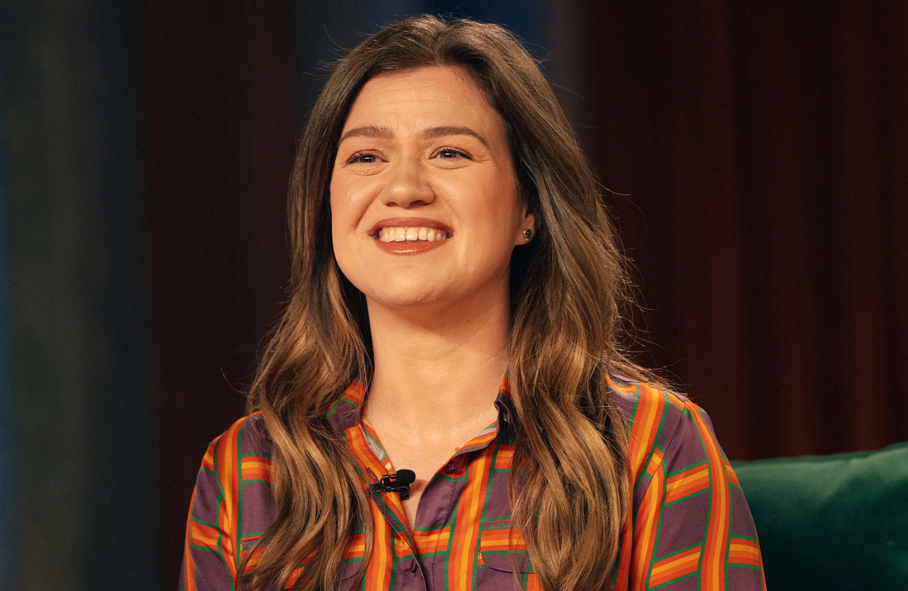 Kelly Clarkson with long brown hair, wearing a colorful striped shirt, smiling.