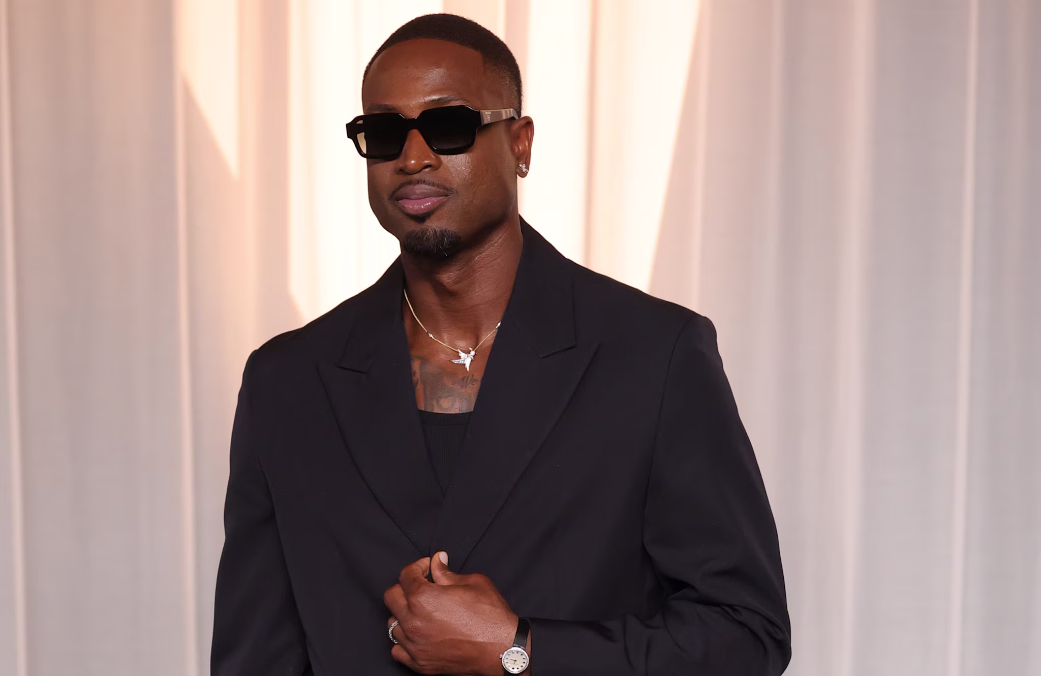 Dwayne Wade wearing sunglasses and a dark suit stands in front of a light curtain, posing confidently with one hand on his jacket.