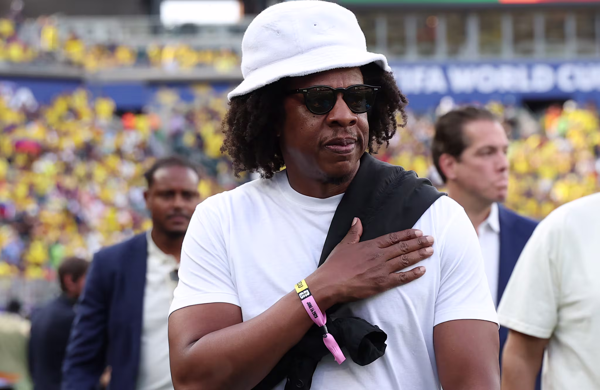 Jay-Z at a sports event, wearing a white bucket hat, sunglasses, and a white t-shirt. He has a hand on his chest.