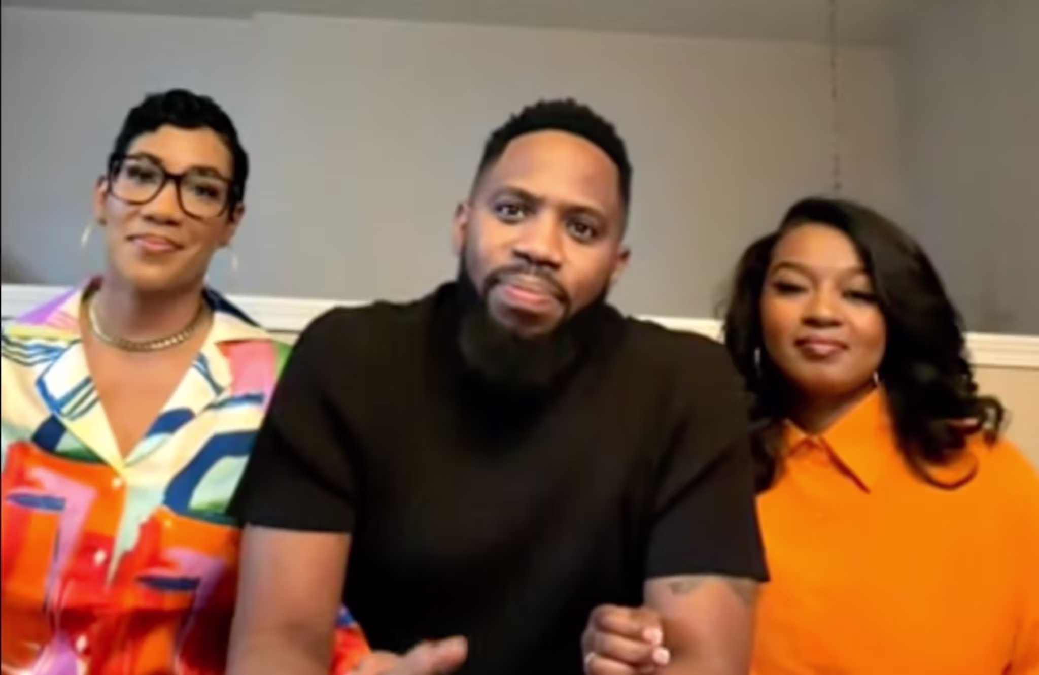 Three people are sitting together, smiling. The person on the left wears glasses and a colorful shirt, the middle person wears a black shirt, and the person on the right wears an orange shirt.