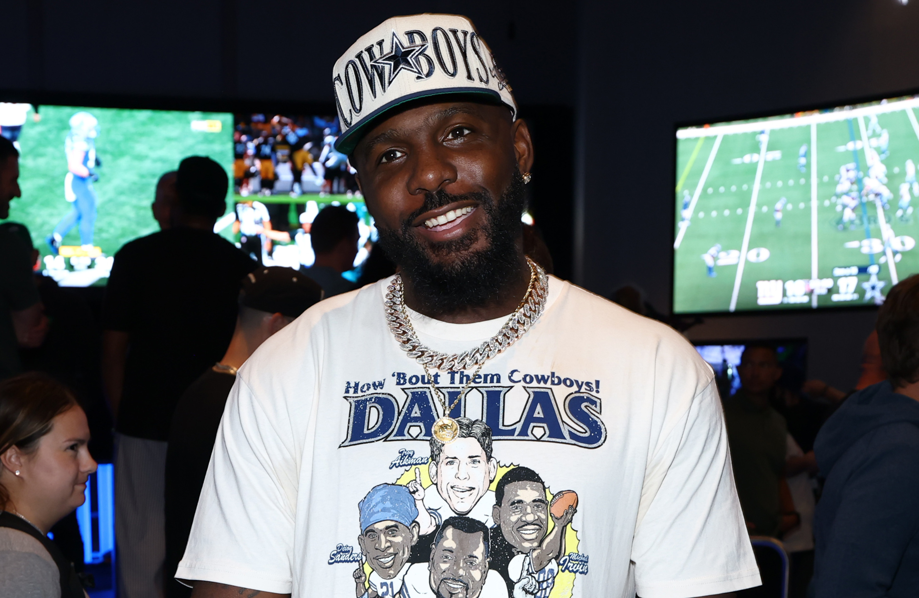Dez Bryant wearing a Dallas Cowboys cap and t-shirt stands in front of TV screens showing football games.