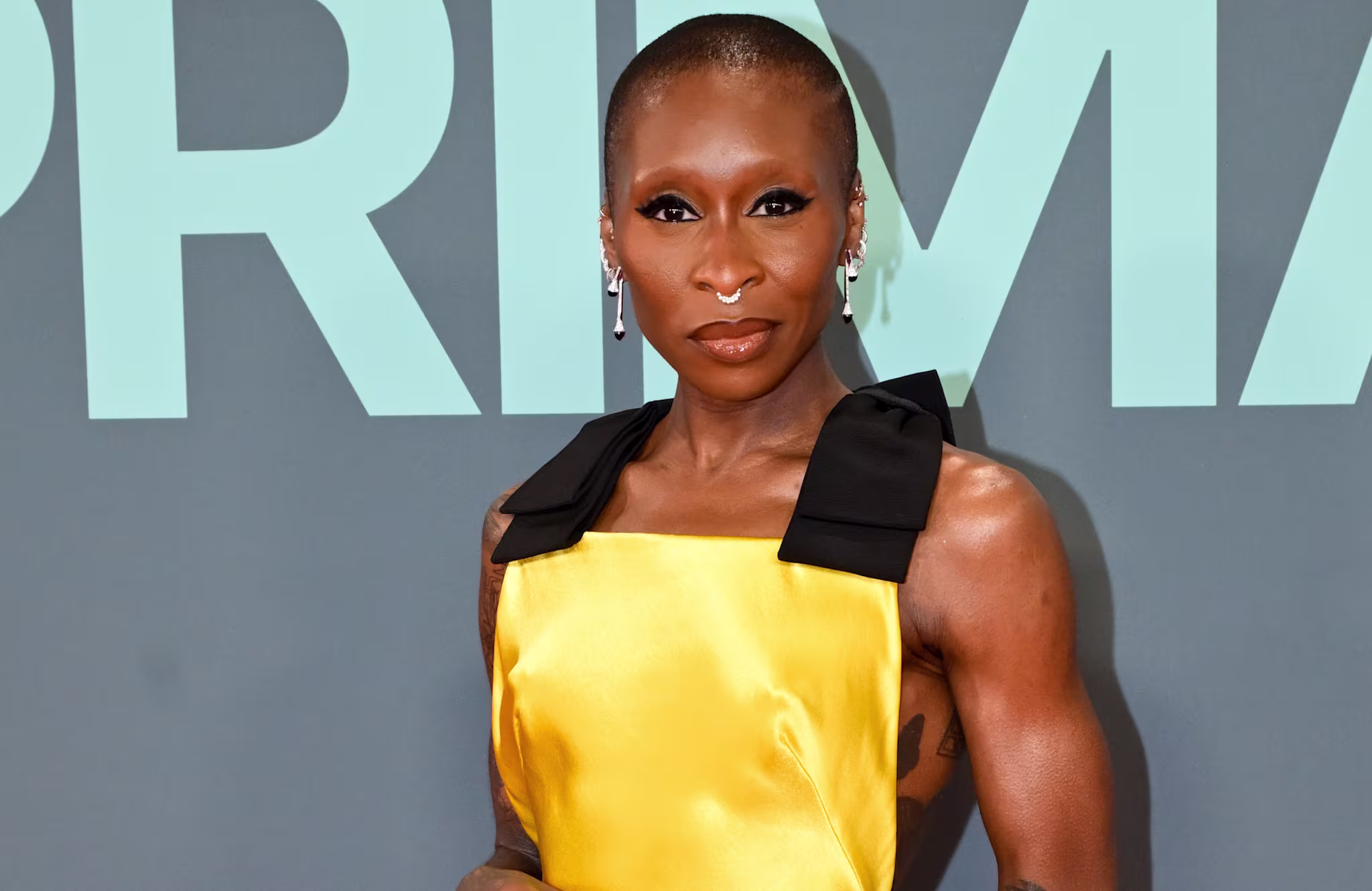 Cynthia Erivo in a yellow dress with black accents, posing against a blue-green background.