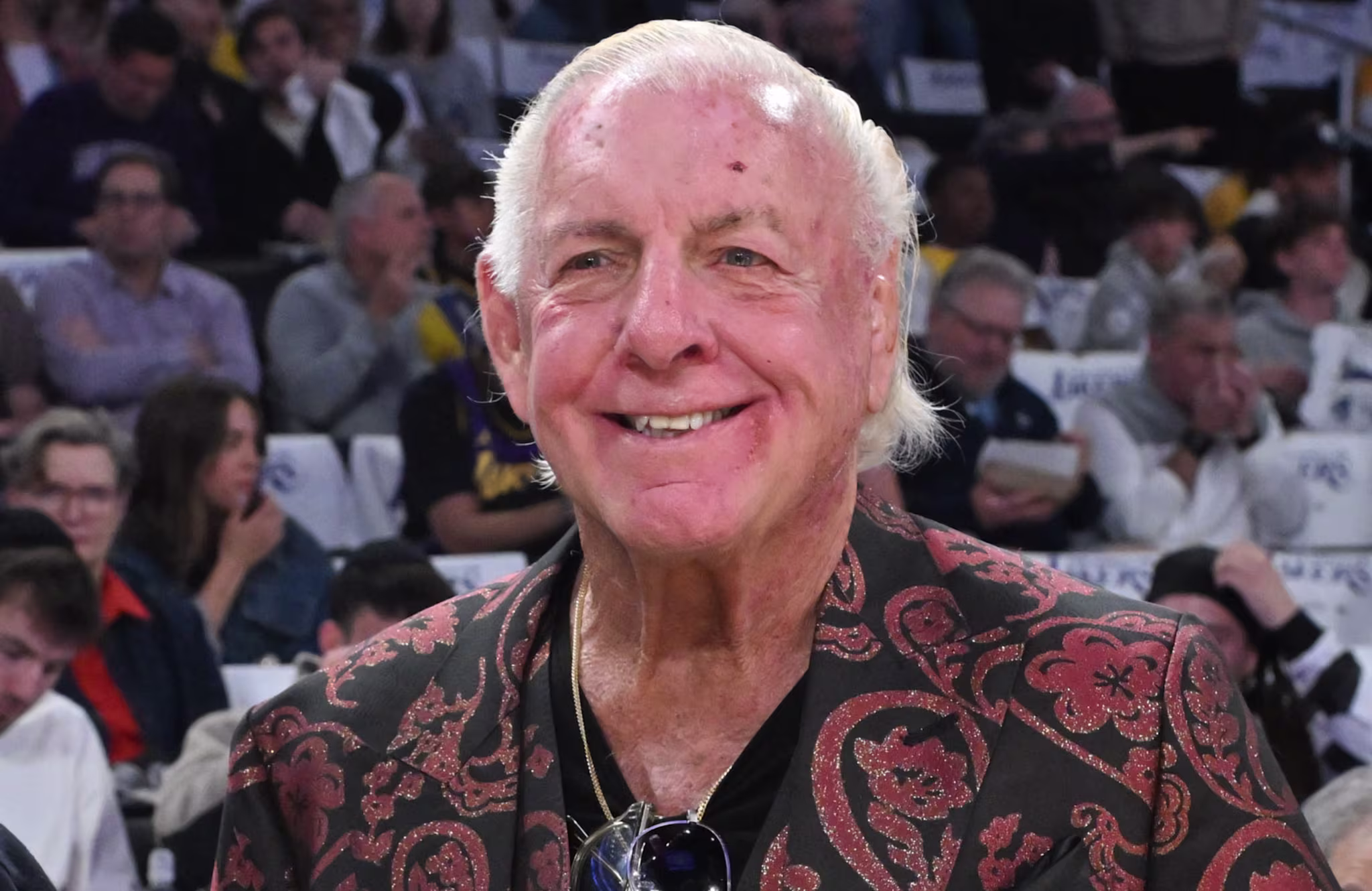 Ric Flair, wearing a patterned jacket, smiles at an event. The background shows a crowd of people seated.