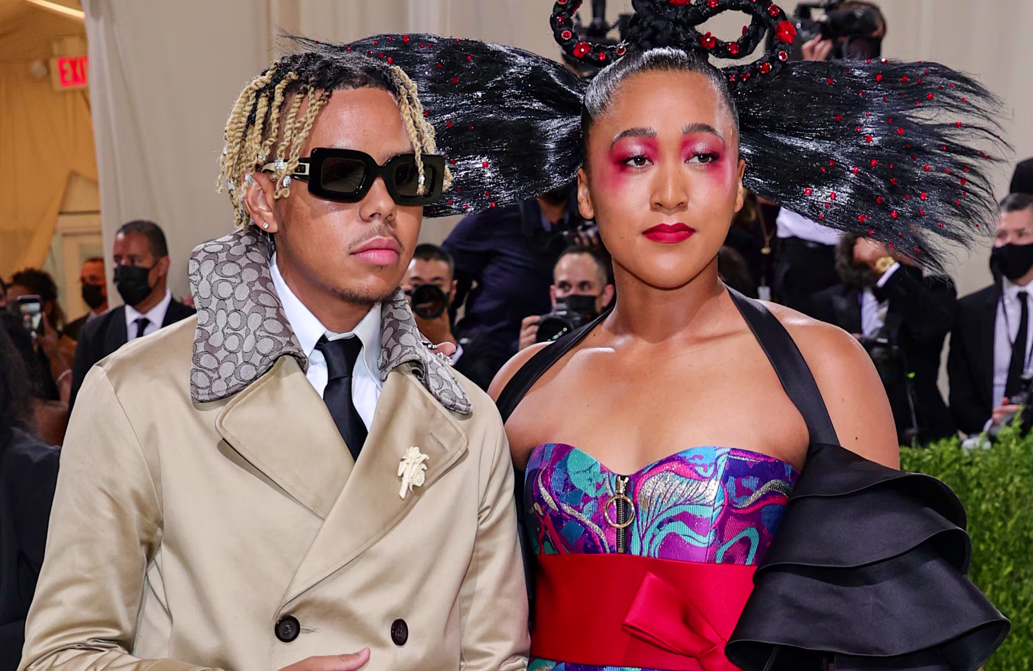 Cordae in a tan coat with sunglasses, and Naomi Osaka in a colorful gown with dramatic hair and makeup, at a formal event.